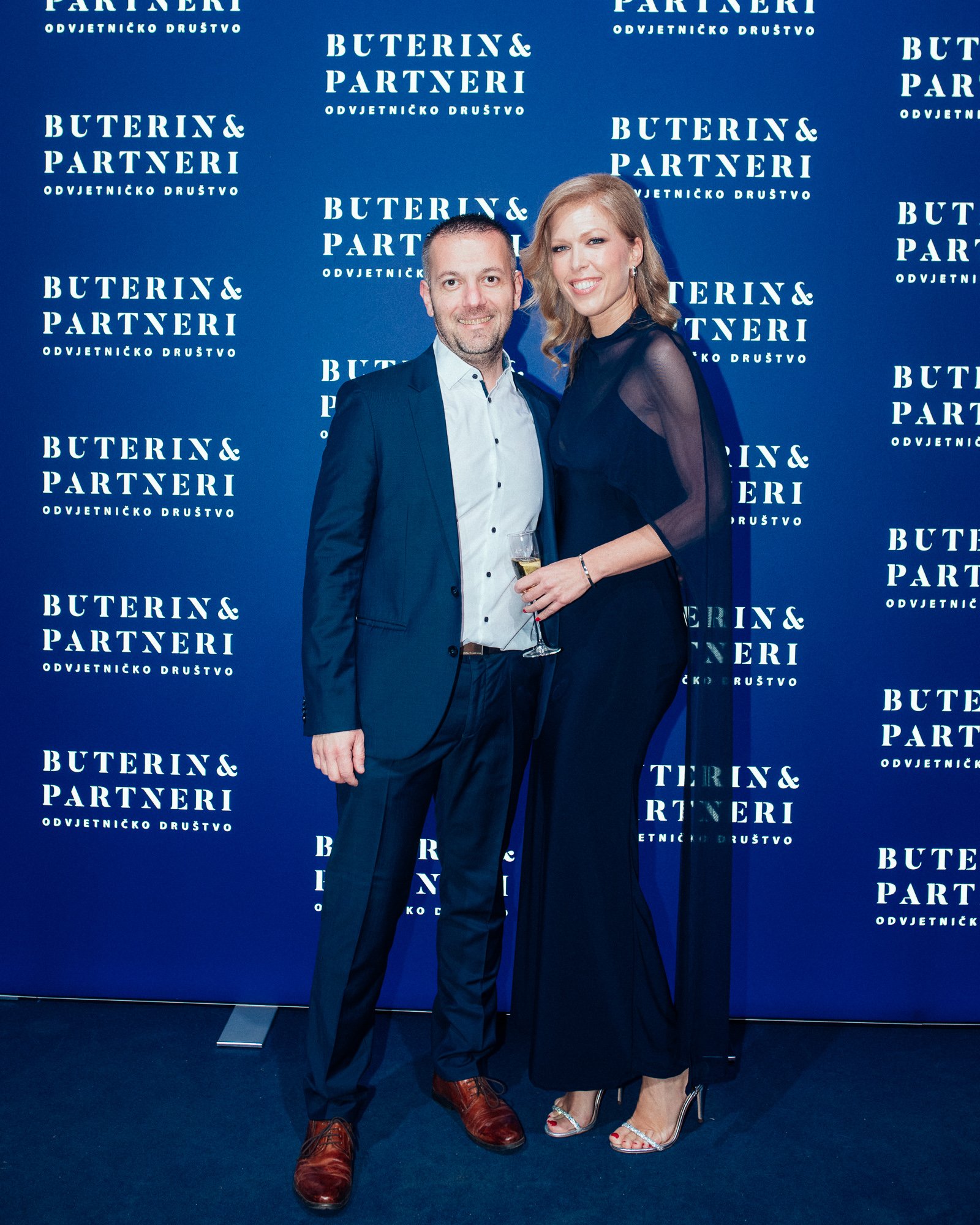 A man and woman in formal attire standing together in front of a blue backdrop with white text that reads 'BUTERIN& PARTNERI ODVJETNIČKO DRUŠTVO'. The man is holding a glass of champagne, and both are smiling.