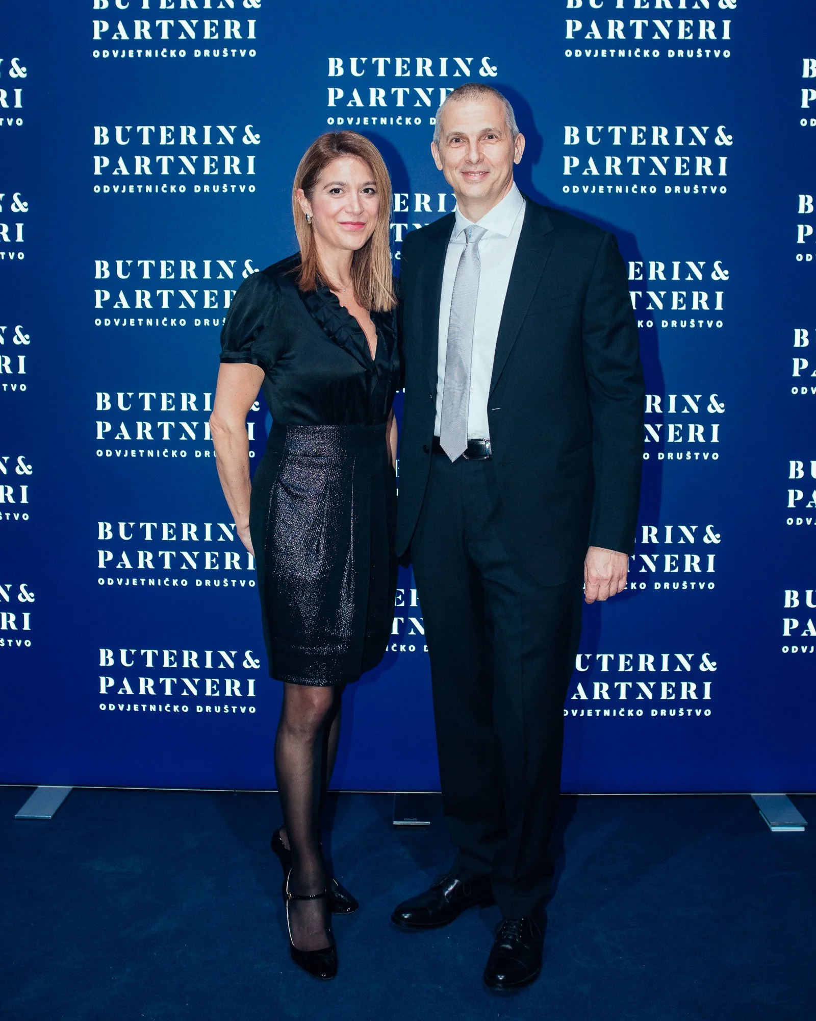 A woman and a man standing in front of a blue backdrop with white text that reads 'BUTERIN & PARTNERI.' The woman is wearing a black blouse, a sparkly skirt, black tights, and black heels. The man is wearing a black suit, white shirt, and a light gra