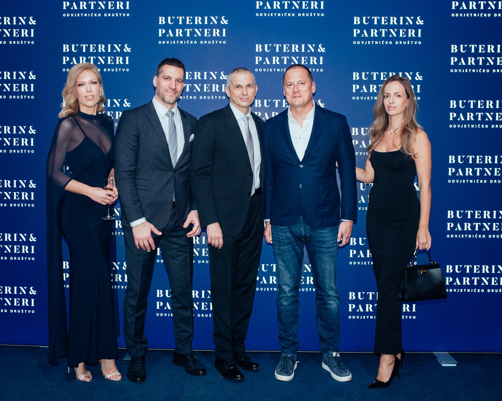 Group of five people dressed formally standing in front of a blue backdrop with the words 'Buterin & Partneri' repeated multiple times, attending an event.