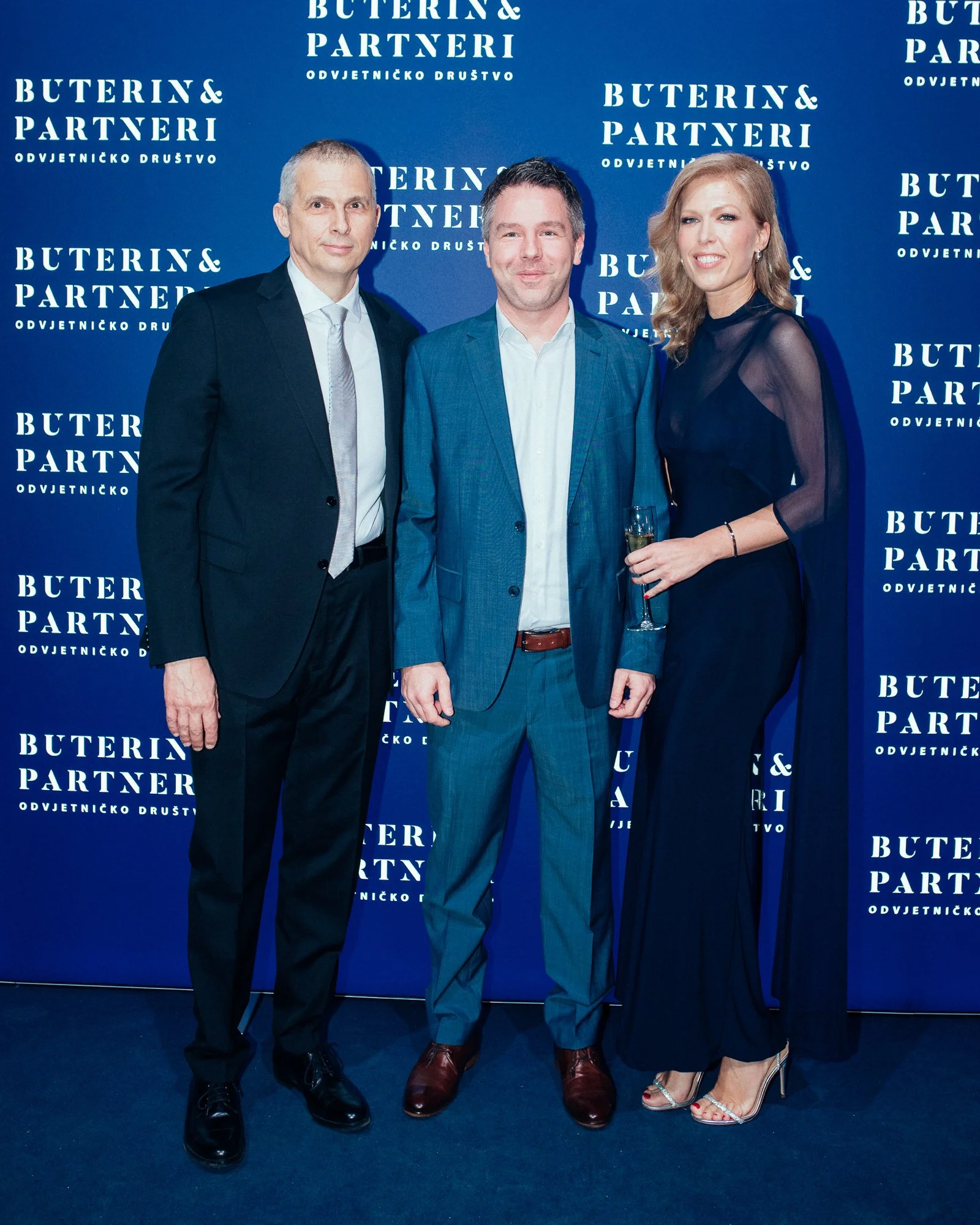 Three people in formal attire standing in front of a blue backdrop with white text that says "Buterin & Partneri, Odvjetničko društvo." The person on the right is holding a glass of champagne.