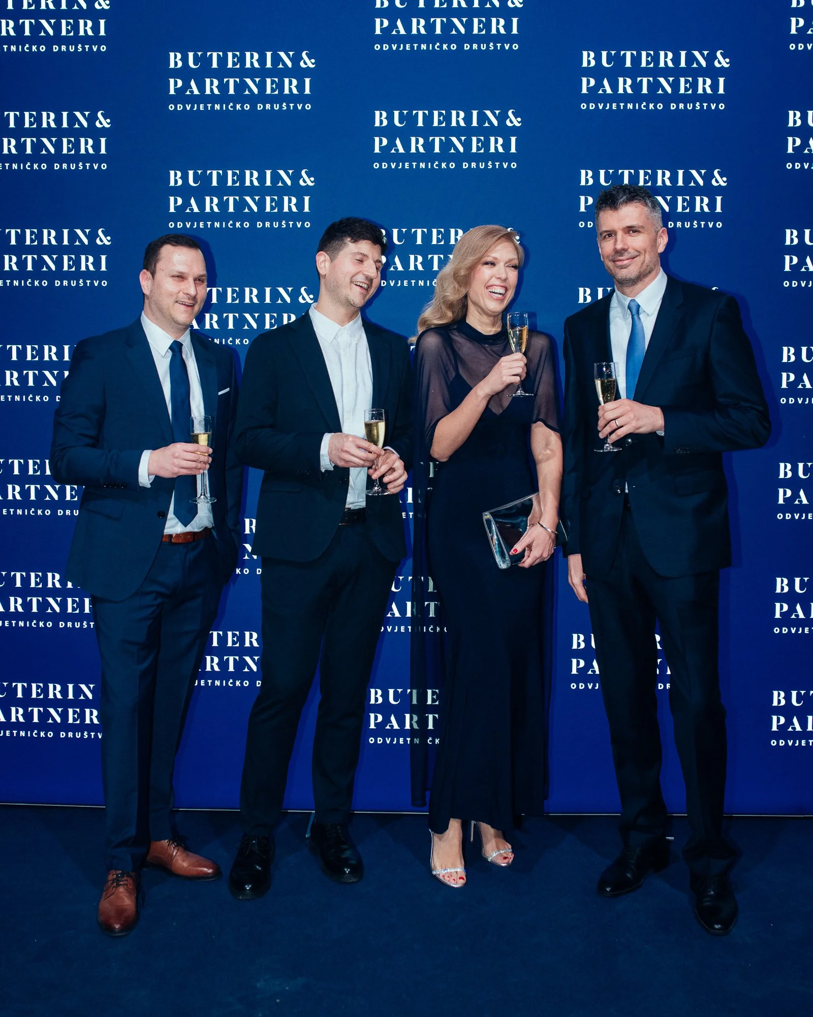 Four people in formal attire, holding champagne glasses, standing in front of a blue backdrop with the text 'Buterin & Partneri' repeated multiple times.