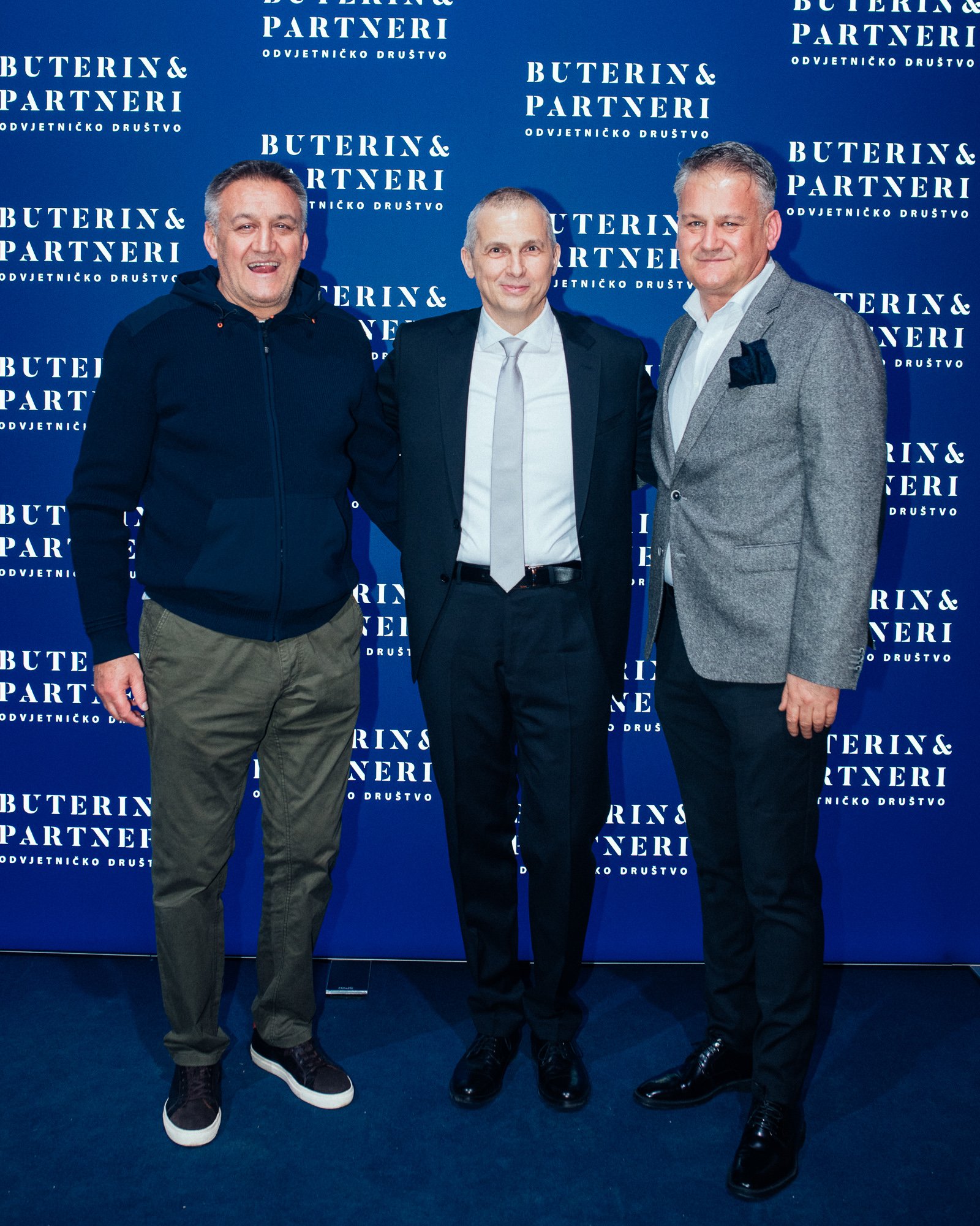 Three men standing in front of a blue backdrop with the text 'BUTERIN& PARTNERI' repeated. They are dressed in formal and casual attire, smiling and posing for a photo.