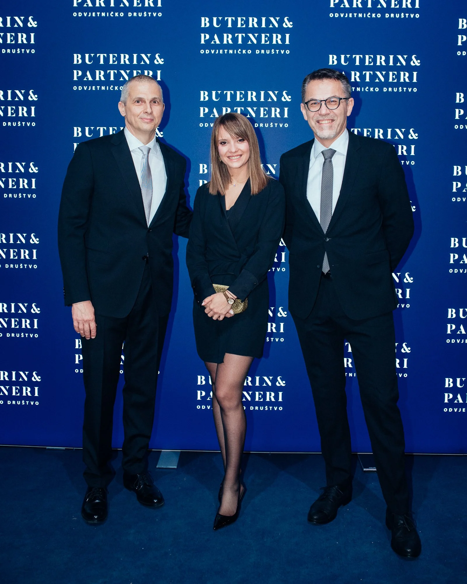 Three people dressed in formal business attire standing in front of a blue backdrop with the repeated text "Buterin & Partneri".