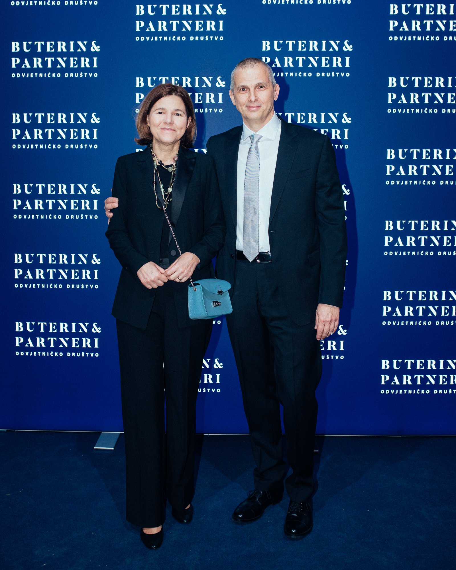 A woman and a man dressed in formal black attire standing together at an event, in front of a blue backdrop with the text 'Buterin & Partneri ODVJETNIČKO DRUŠTVO' repeated multiple times.