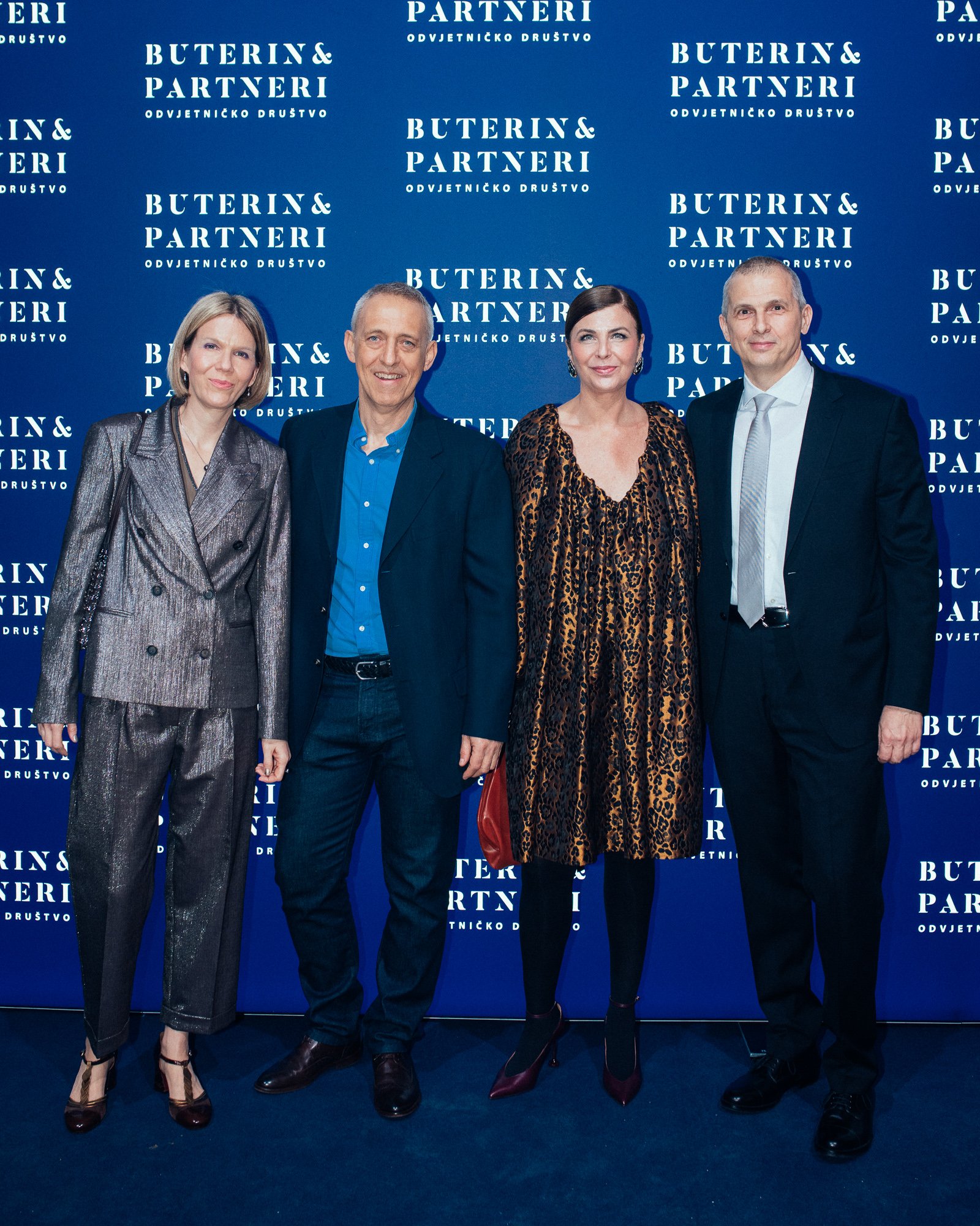 Four people standing in front of a blue background with the logo 'Buterin & Partneri' and additional text in a foreign language, dressed in formal attire, for a professional or social event.