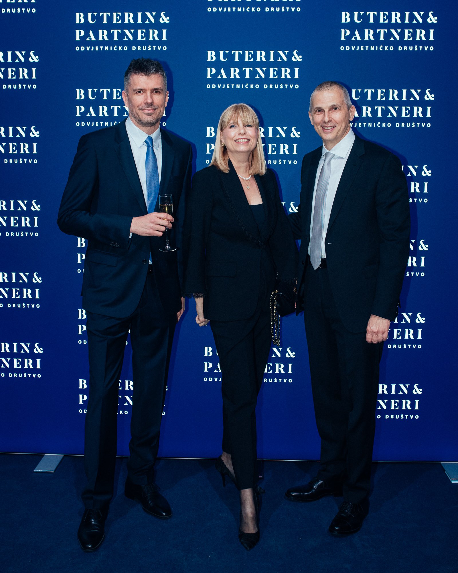 Three people in formal attire standing in front of a blue backdrop with the words 'Buterin & Partneri Odvjetničko Društvo' repeated, one person holding a glass of champagne.