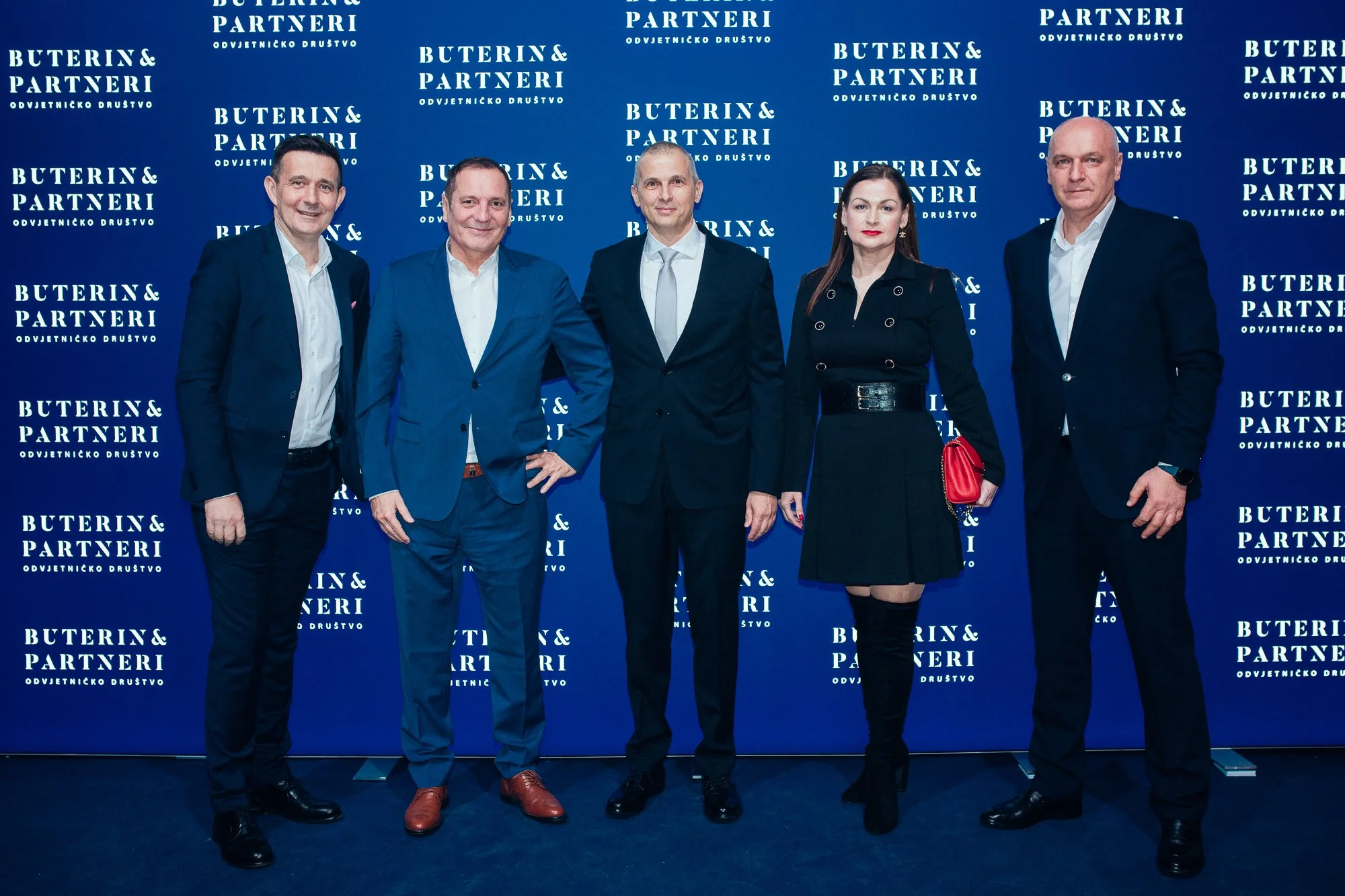 Five people standing in front of a blue backdrop with white text reading 'Buterin & Partneri.' They are dressed in formal business attire, with four men in suits and one woman in a black dress and boots. The group is smiling and facing the camera.