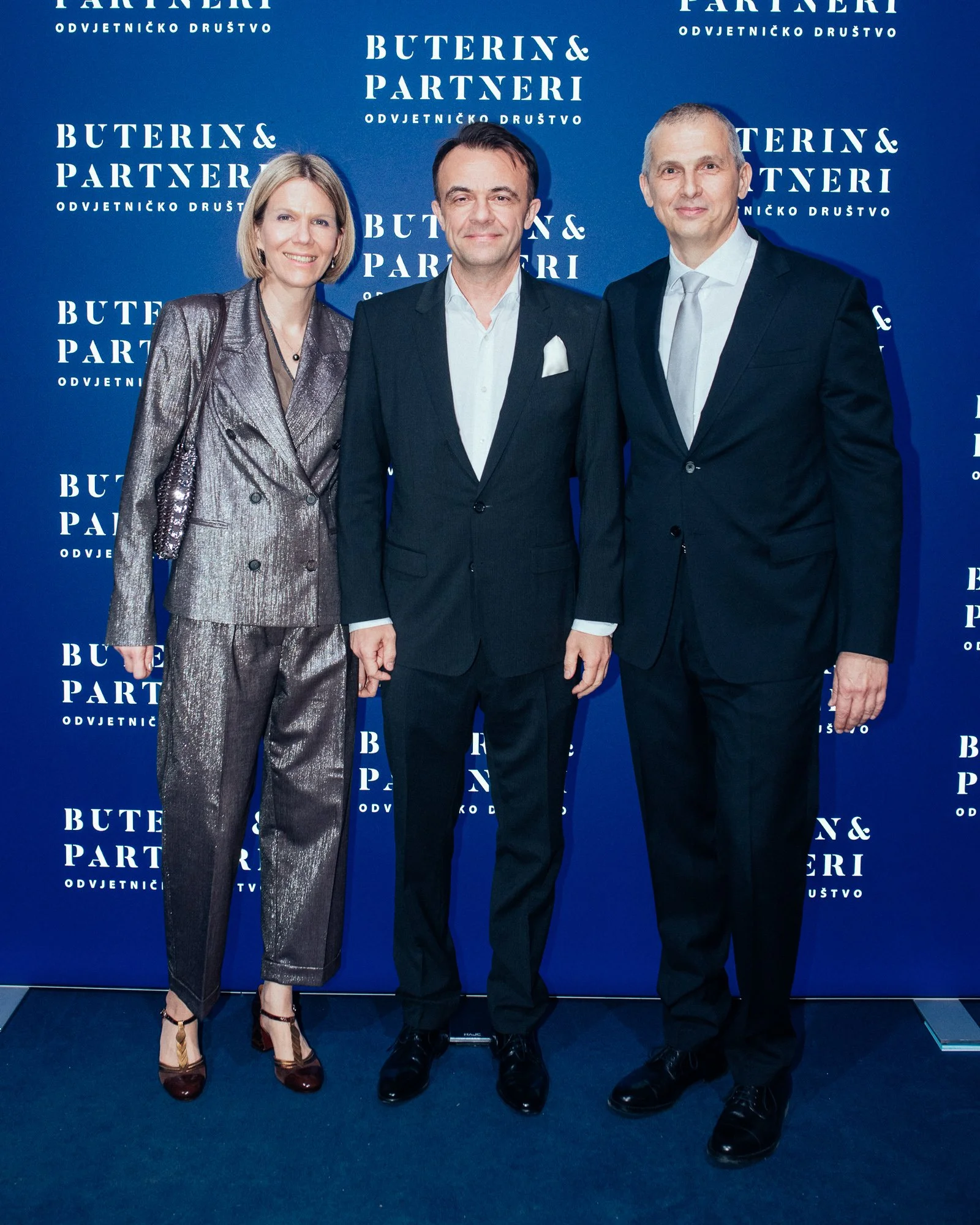 Three people dressed in formal attire standing in front of a blue backdrop with the words 'BUTERIN & PARTNERI ODVJETNICKO DRUŠTVO' printed repeatedly.