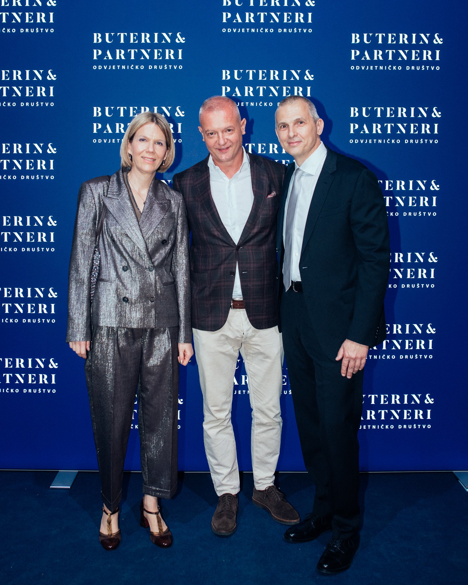 Three people standing in front of a blue backdrop with white text that reads 'BUTERIN & PARTNERI ODVJETNIČKO DRUŠTVO.' The woman on the left is wearing a shiny silver suit and dark heels, the man in the middle is dressed in a dark plaid blazer, white