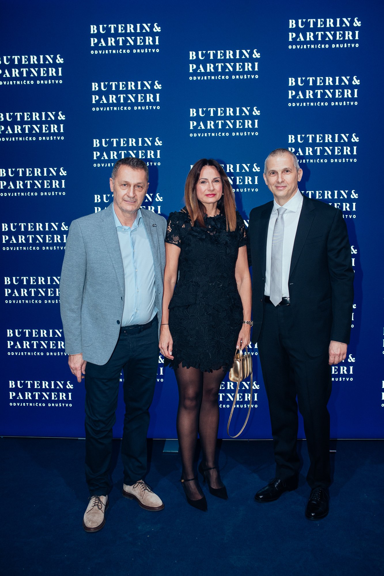 Three people, two men and a woman, standing in front of a blue backdrop with white text that reads 'Buterin & Partneri Odvjetničko Društvo'. The man on the left is wearing a light gray blazer, blue shirt, and dark pants. The woman in the middle is dr