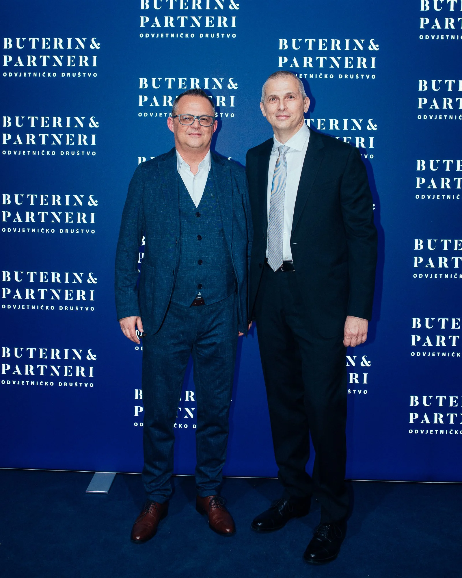 Two men in suits standing in front of a blue backdrop with white text that reads 'Buterin & Partneri Odvjetničko društvo'. The man on the left is wearing glasses, a blue checked suit, white shirt, and brown shoes. The man on the right is bald, in a b