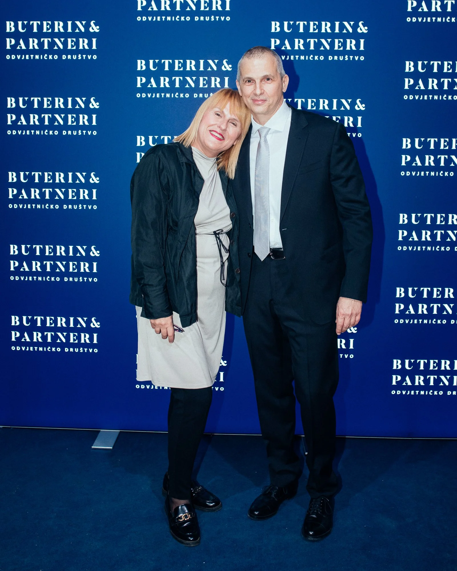 A woman and a man standing together at a formal event, posing for a photo in front of a blue backdrop with white text that reads 'Buterin & Partneri' and 'Odvjetničko Društvo'. The woman is smiling, leaning her head on the man's shoulder, and the man