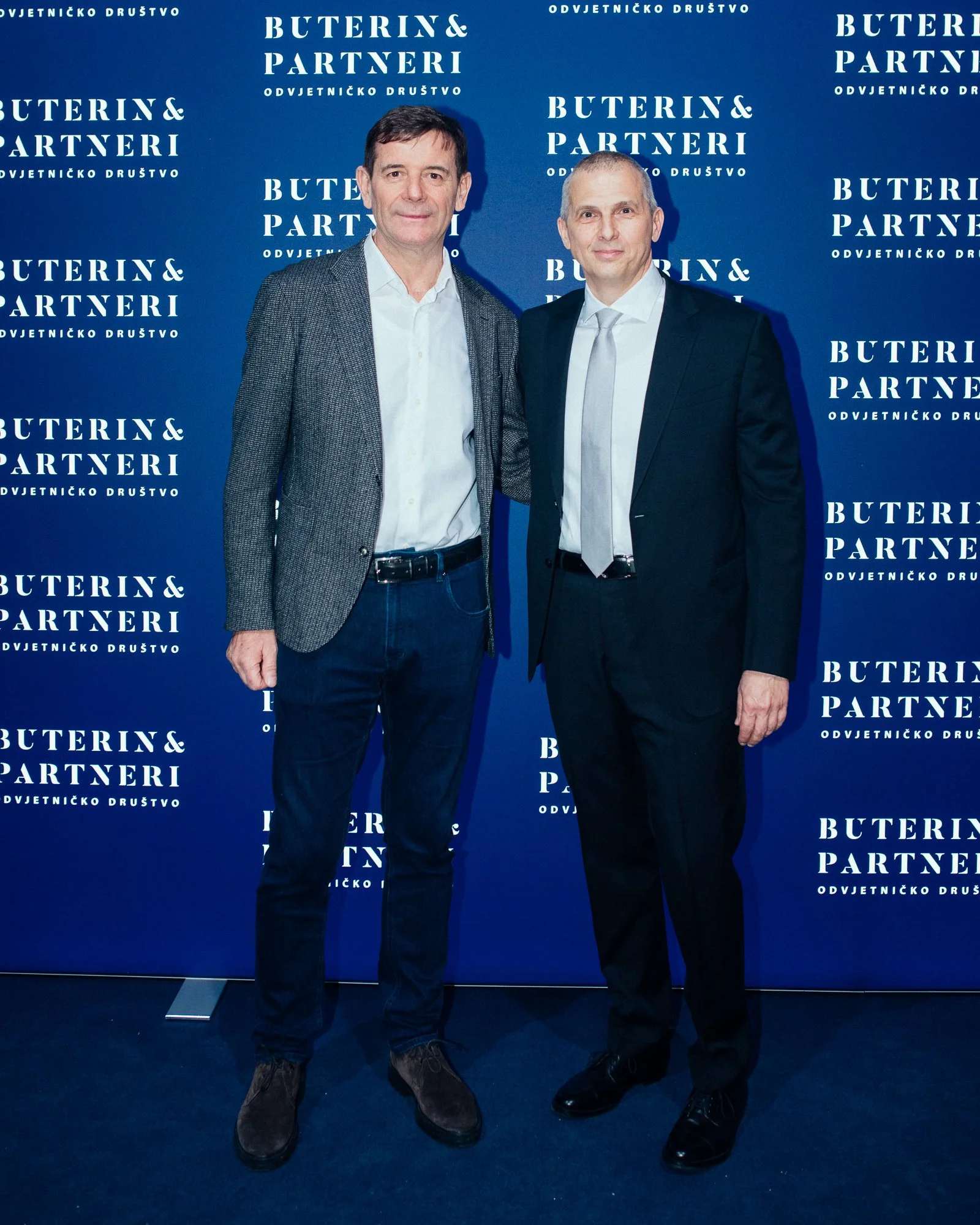 Two men in business attire standing in front of a blue backdrop with white text that reads 'BUTERIN& PARTNERI' and 'ODVJETNIČKO DRUŠTVO'. One man is wearing a grey blazer, white shirt, and dark jeans, while the other is wearing a black suit with a wh