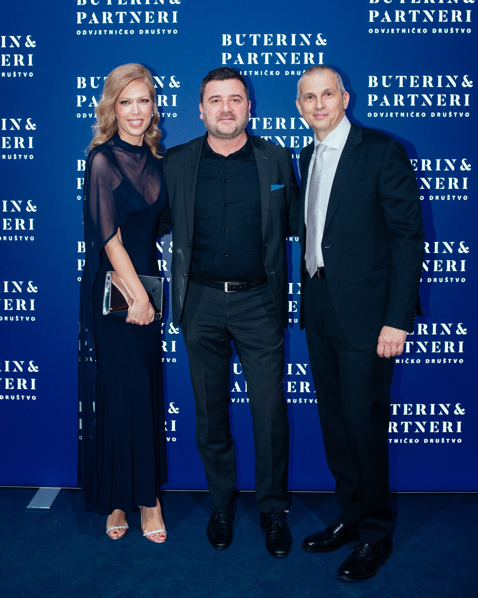 Three people standing together at a formal event in front of a blue backdrop with the text 'Buterin & Partneri' and 'Odvjetničko društvo'. The woman on the left is wearing a navy blue dress with sheer sleeves, holding a clutch bag, and wearing open-t