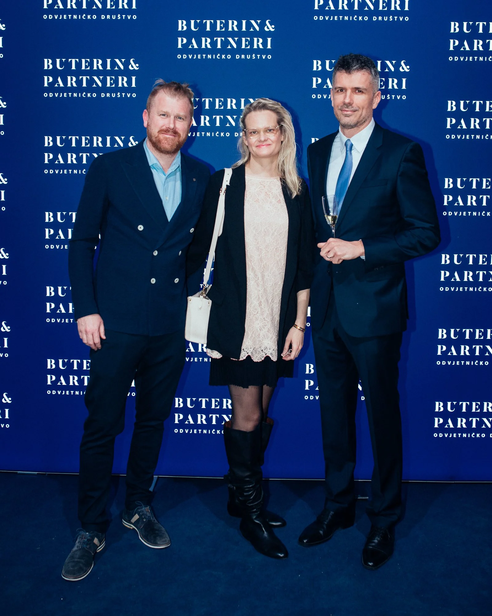 Three people standing together at a formal event, with a blue backdrop that has the text 'Buterin & Partneri' repeatedly printed on it. The person on the left is a man with reddish hair and beard, dressed in a dark suit and light blue shirt. The woma