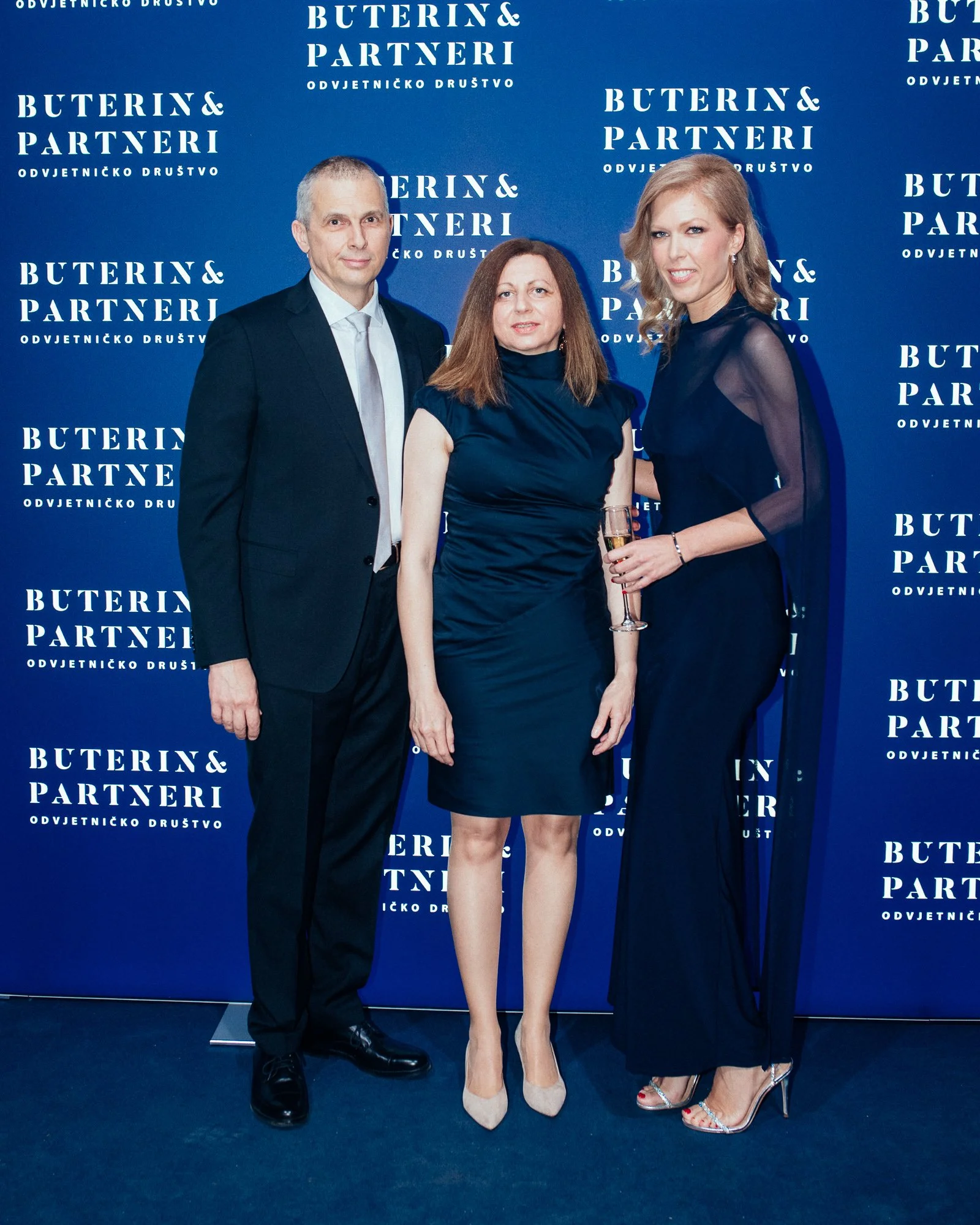 Three people dressed in formal attire standing in front of a blue backdrop with white text that reads "Buterin & Partneri Odvjetničko Društvo." The woman on the right is holding a champagne glass. They are posing for a photo at a formal event.