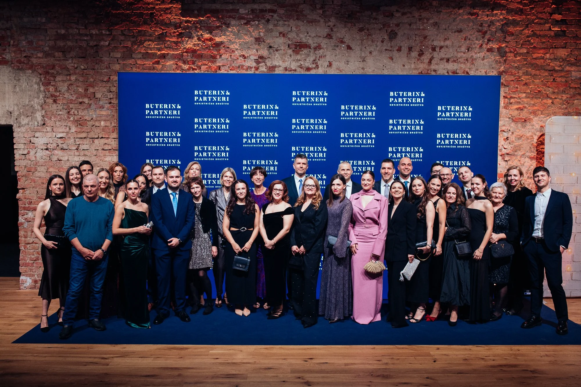 A large group of people dressed in formal attire standing in front of a blue backdrop with white text that reads 'BUTERIN & PARTNERI ODVJETNICKO DRUSTVO'. They are posing for a photo at a professional or corporate event in an indoor venue with expose