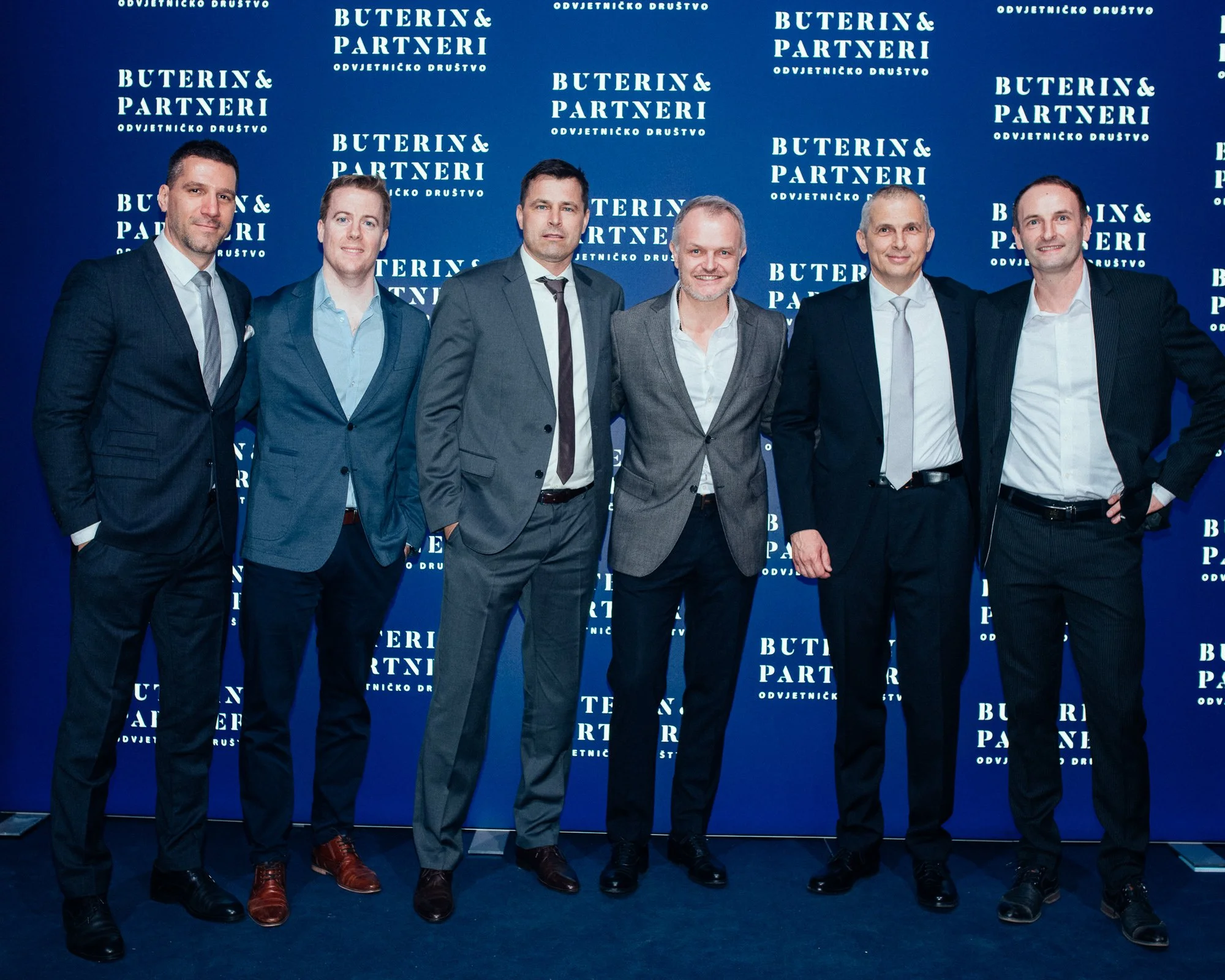 Seven men in business suits standing in front of a blue backdrop with the text 'Buterin & Partneri' repeated in white, posing for a photo at a professional event.