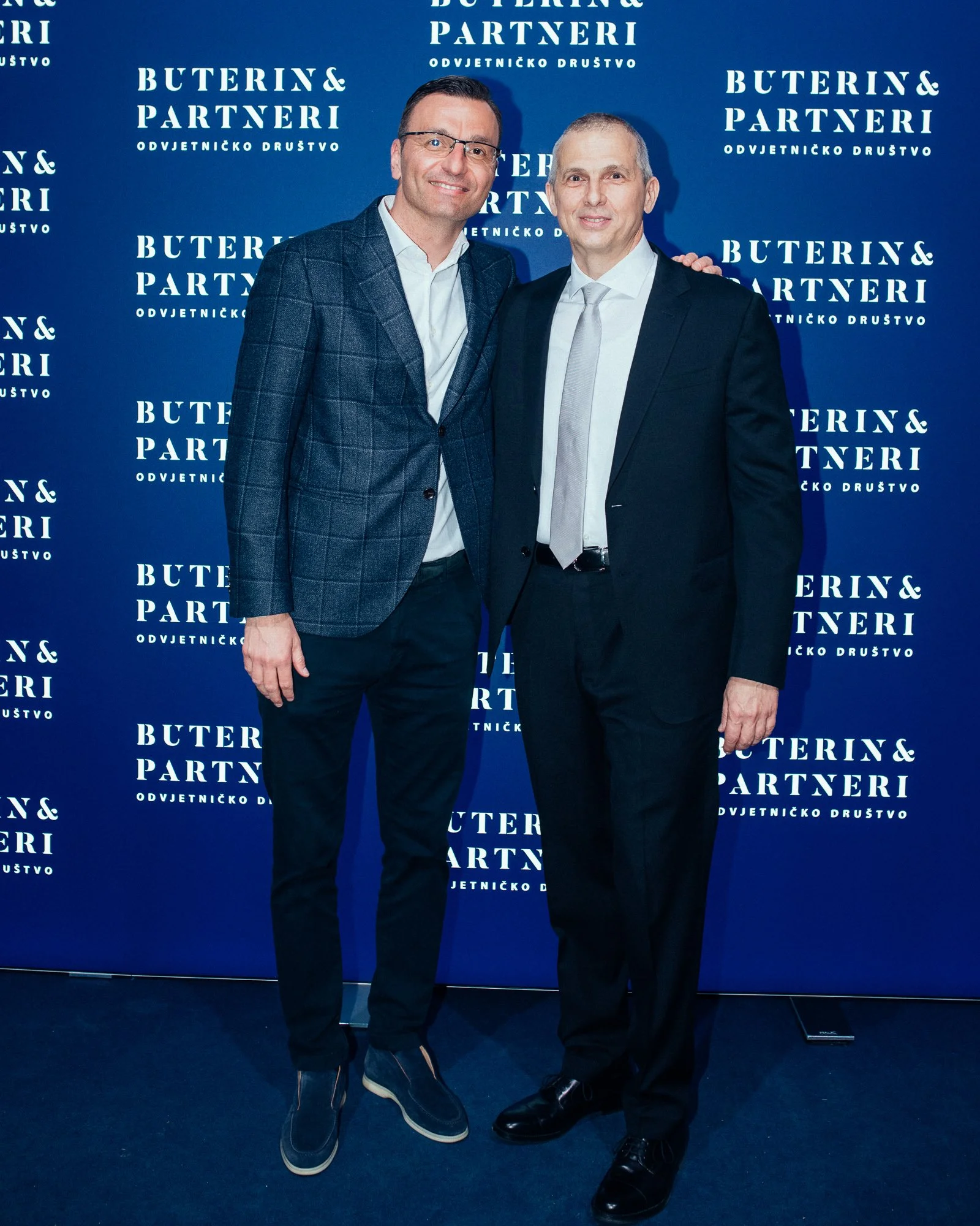 Two men in formal suits posing together at an event, standing in front of a blue backdrop with white text that reads "Buterin & Partneri." They are smiling, with one man touching the other's shoulder.