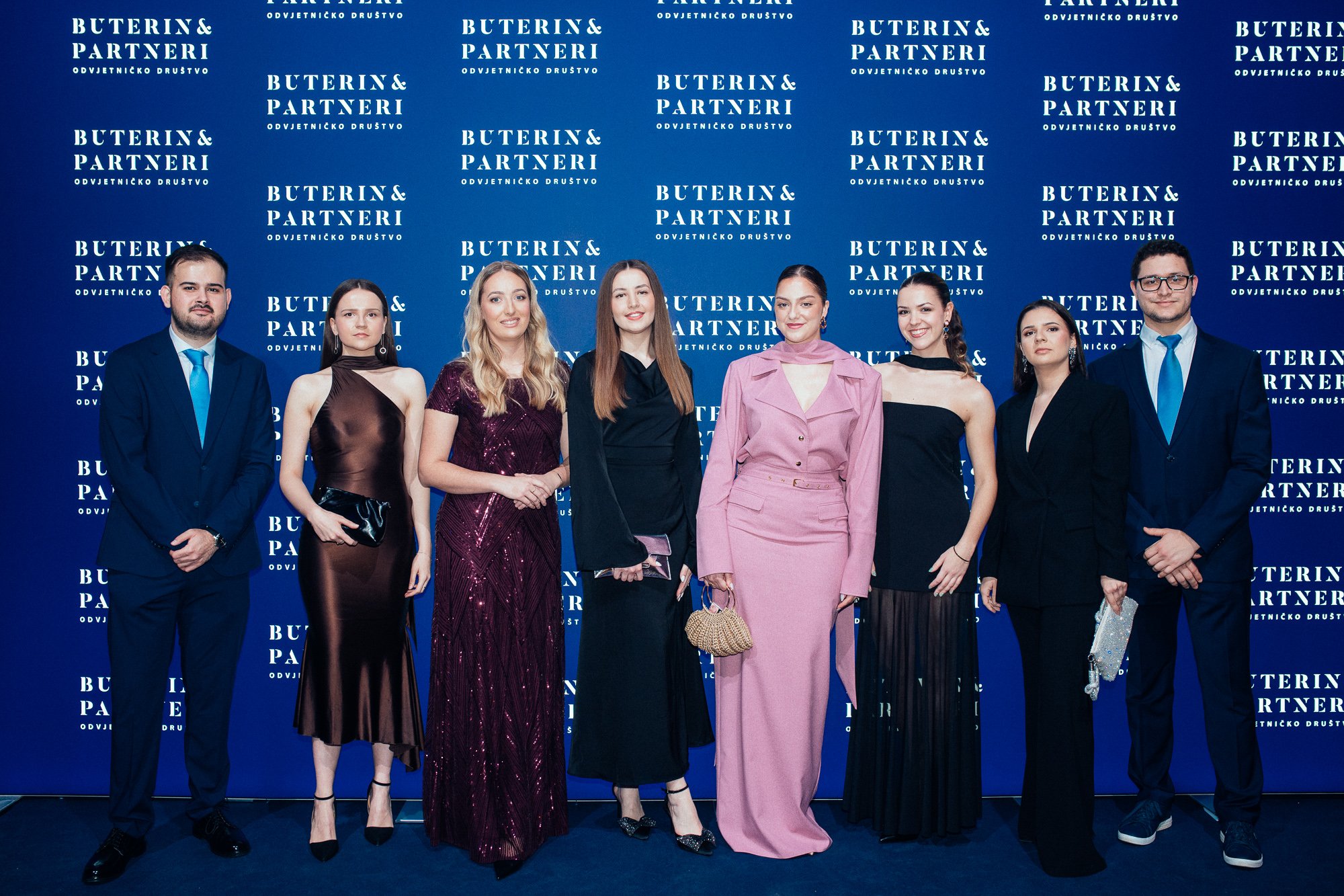 A group of nine young adults, dressed in formal attire, standing in front of a blue backdrop with white text that reads 'Buterin & Partneri Odvjetničko Drustvo.' The group includes five women and four men.