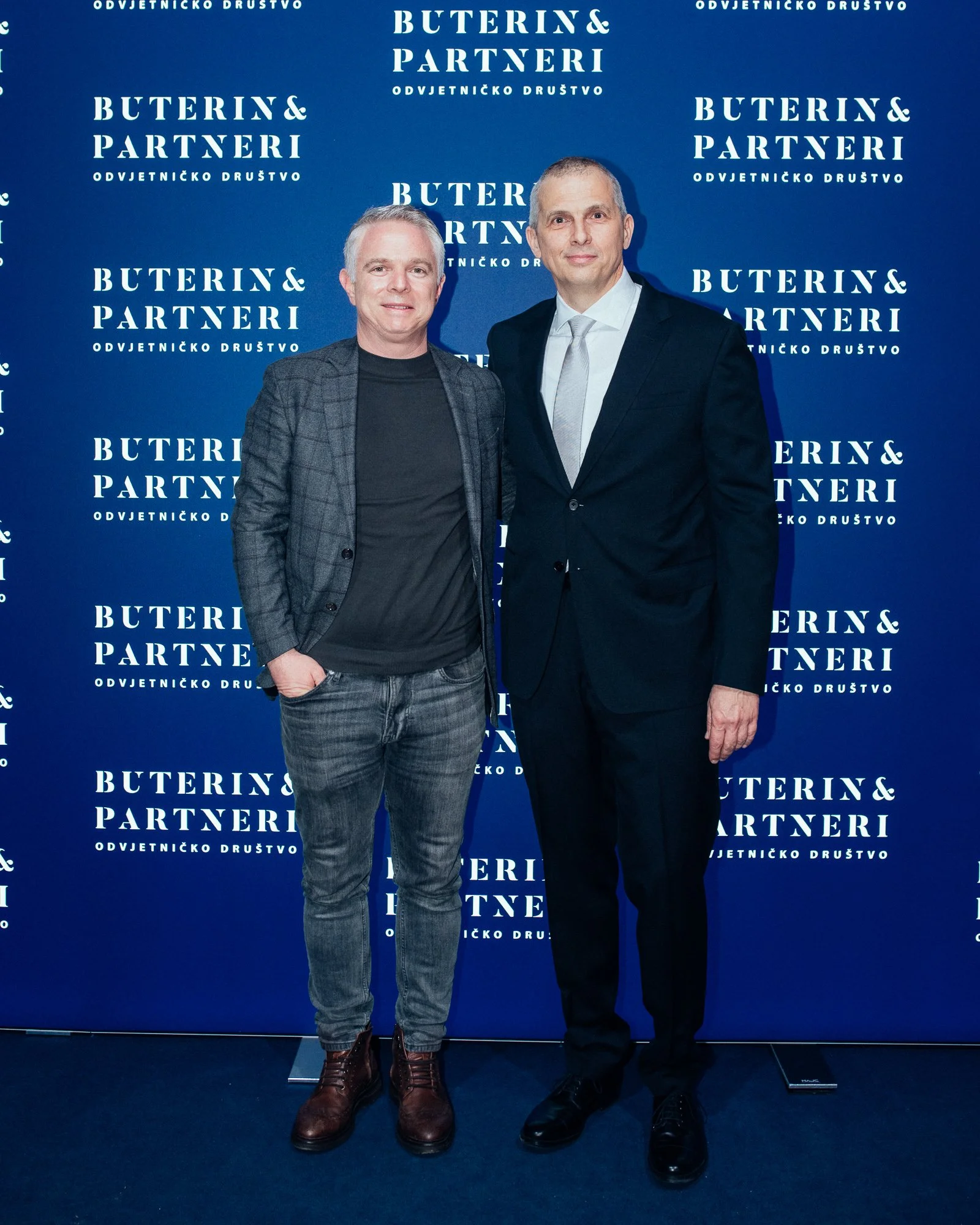 Two men standing in front of a blue backdrop with the text "Buterin & Partneri" and additional text in a foreign language, dressed in business attire.