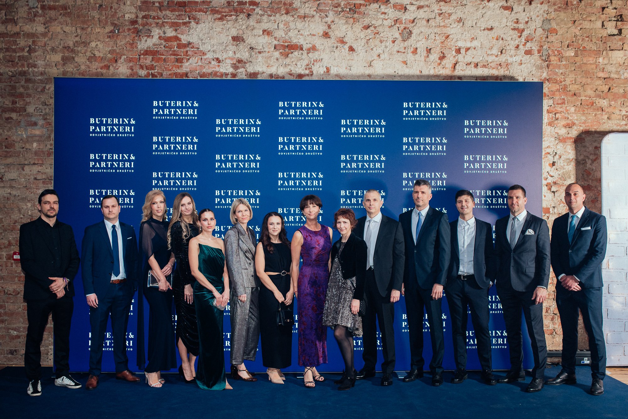 Group of fifteen people in formal attire standing in front of a blue backdrop with a brick wall behind it, at an event branded 'Buterin & Partneri'.