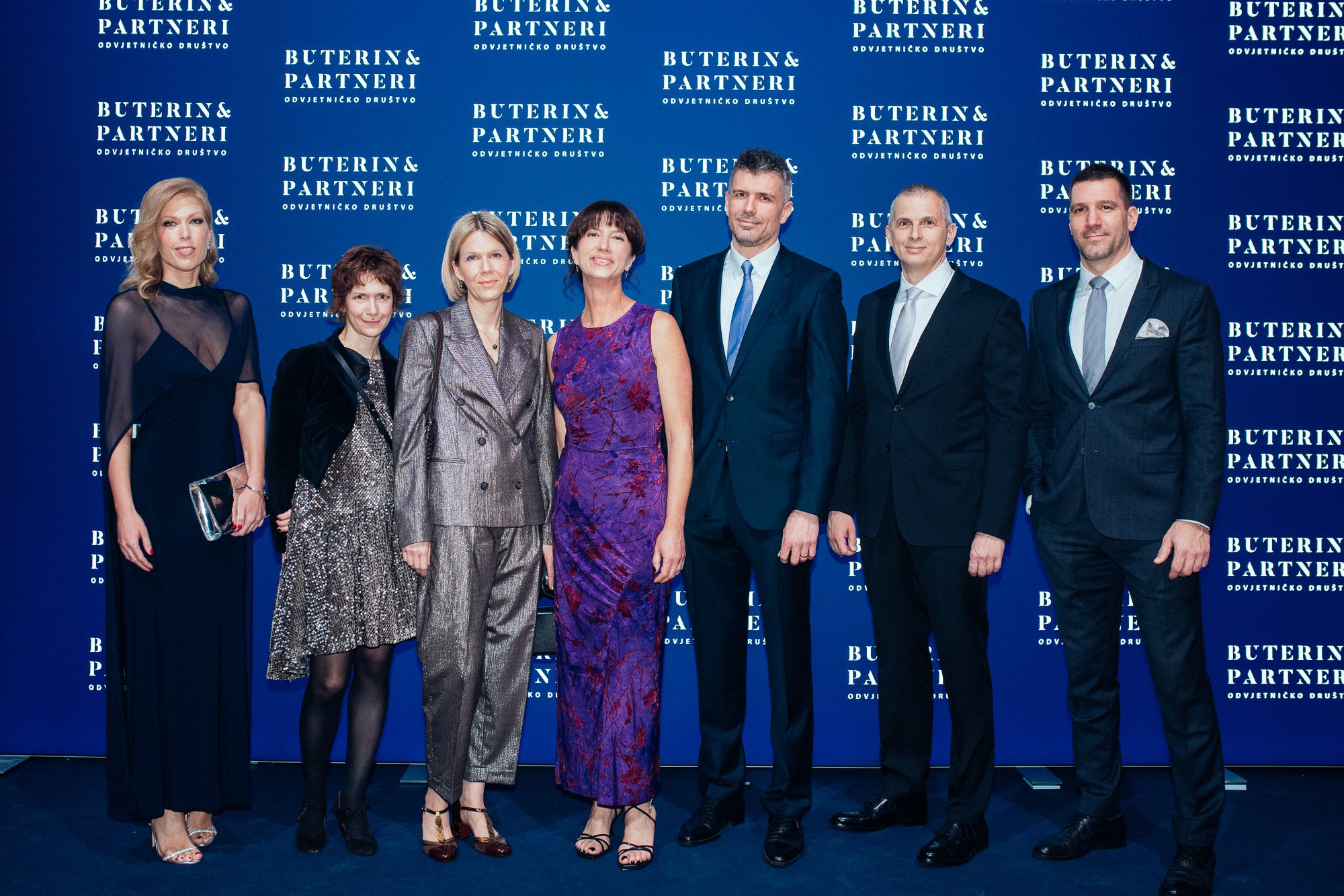 Group of seven people in formal attire standing in front of a blue backdrop with white text that reads 'Buterin & Partneri Odvjetničko društvo'.