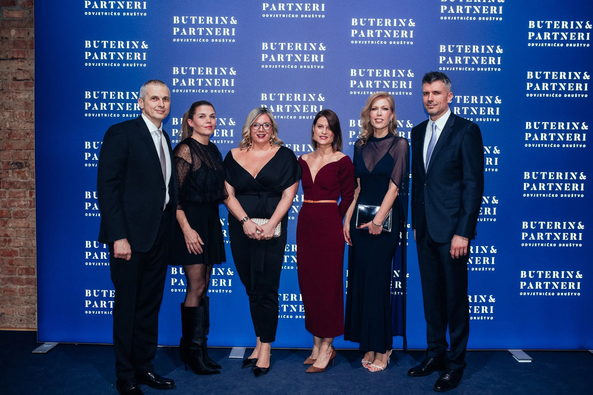 Group of six people dressed in formal attire standing in front of a blue backdrop with a logo that reads "Buterin & Partneri" multiple times, smiling at the camera.