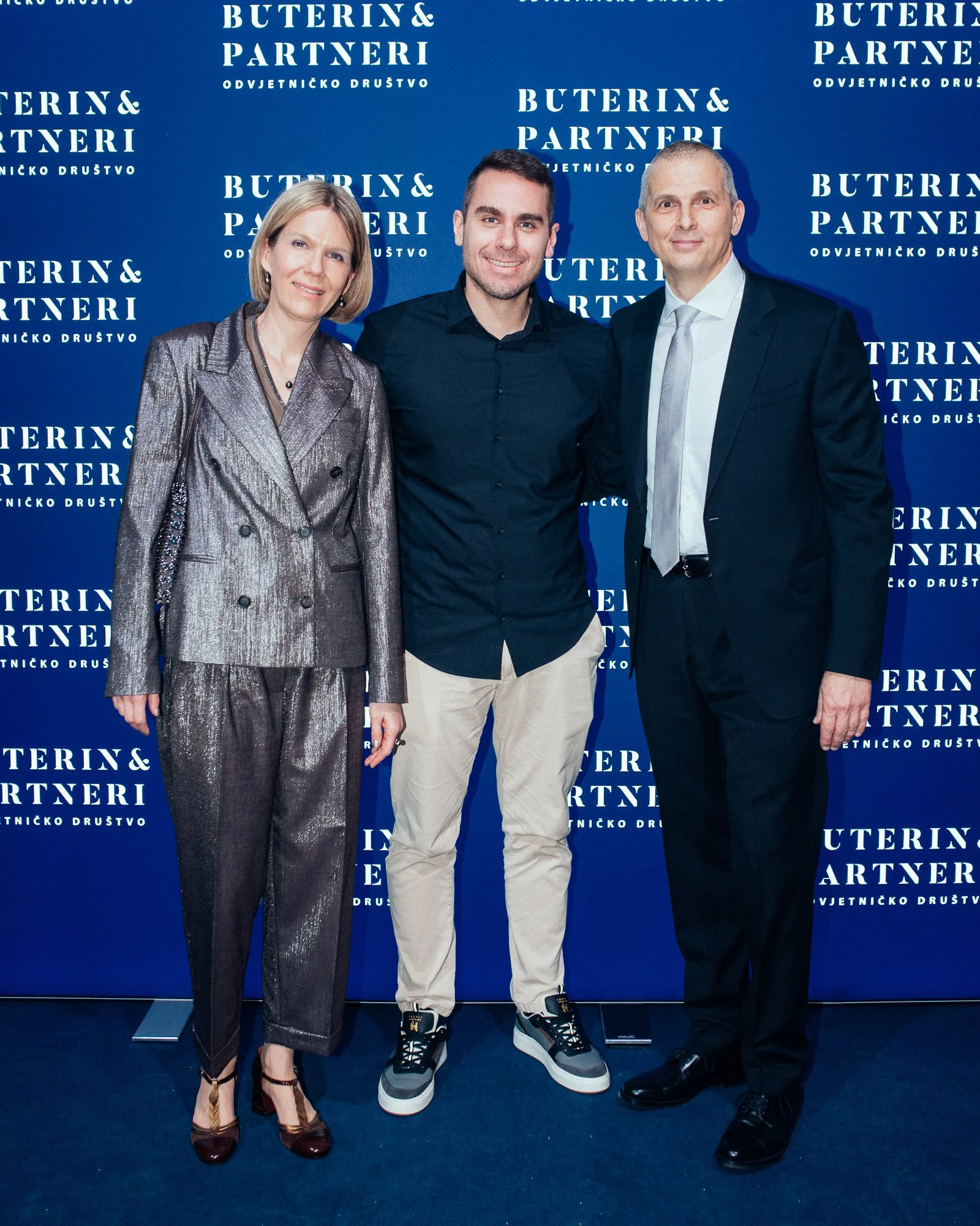 Three people standing in front of a blue backdrop with white text that reads 'Buterin & Partneri' and 'Odvjetničko društvo.' The person on the left is a woman wearing a shiny silver suit with matching pants and high heels. The person in the middle is