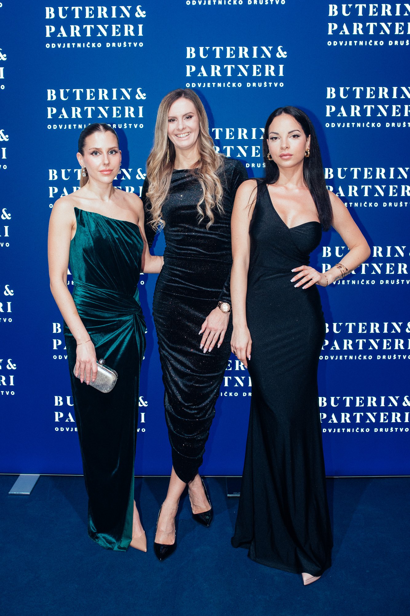 Three women in elegant evening dresses standing in front of a blue backdrop with white text that reads 'BUTERIN & PARTNERI ODVJETNICKO DRUSTVO'.