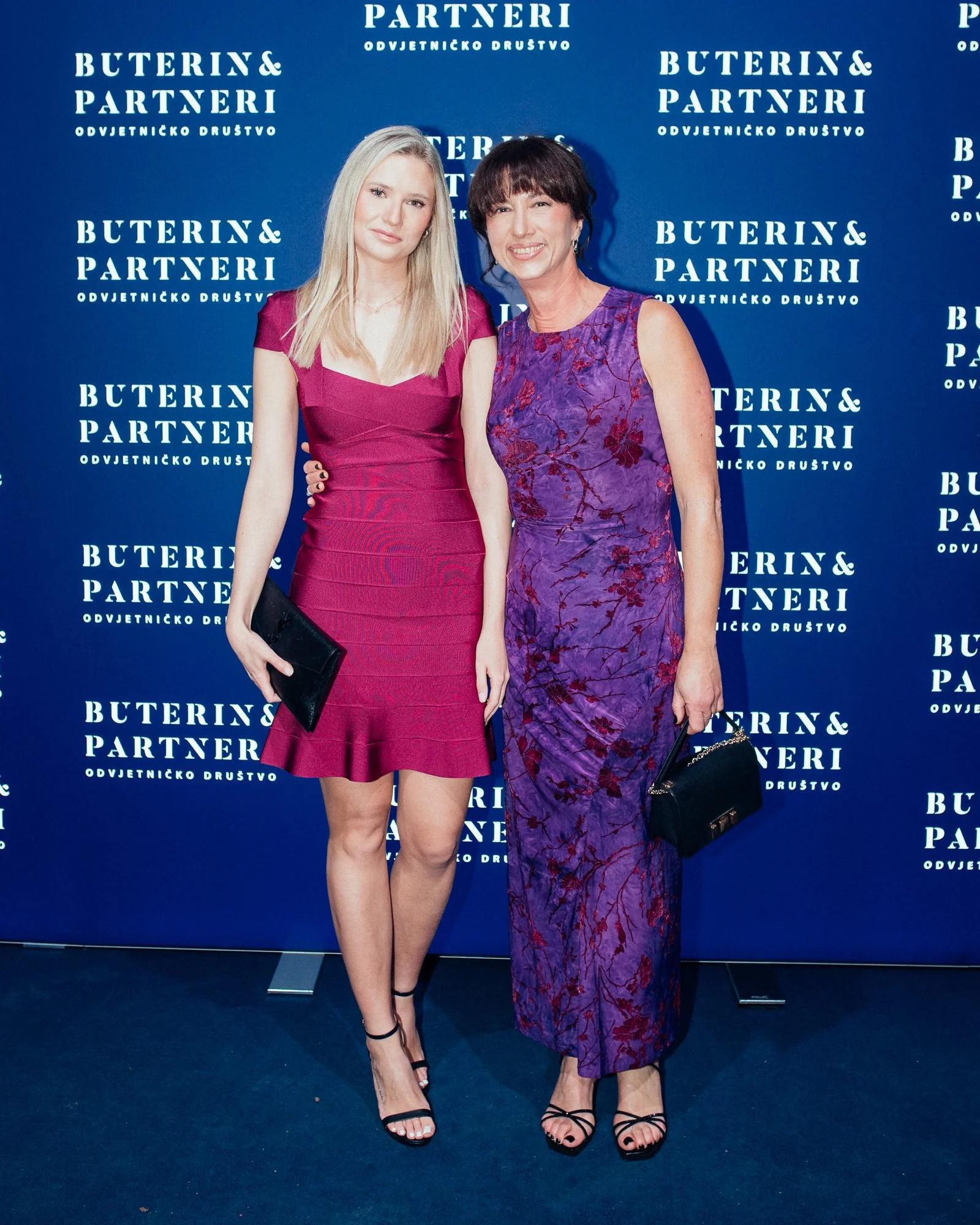 Two women in formal dresses standing in front of a blue backdrop with white text that reads 'Buterin & Partnery Odvjetničko Društvo.'