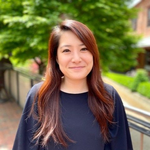 A smiling woman with long reddish-brown hair standing outdoors on a sidewalk with green trees and a black railing in the background.