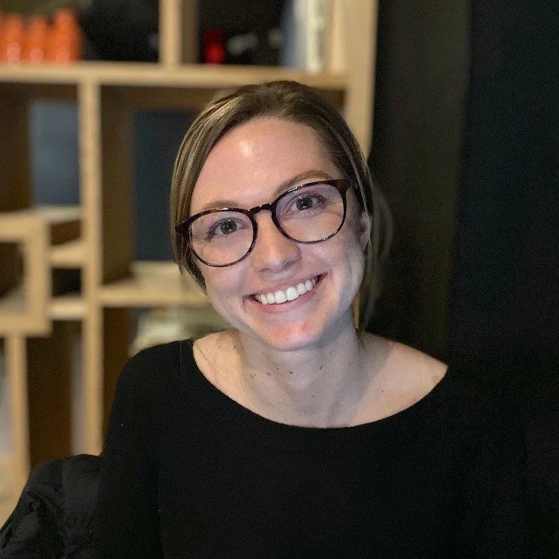 A woman with short brown hair and glasses smiling at the camera in an indoor setting with wooden shelves in the background.