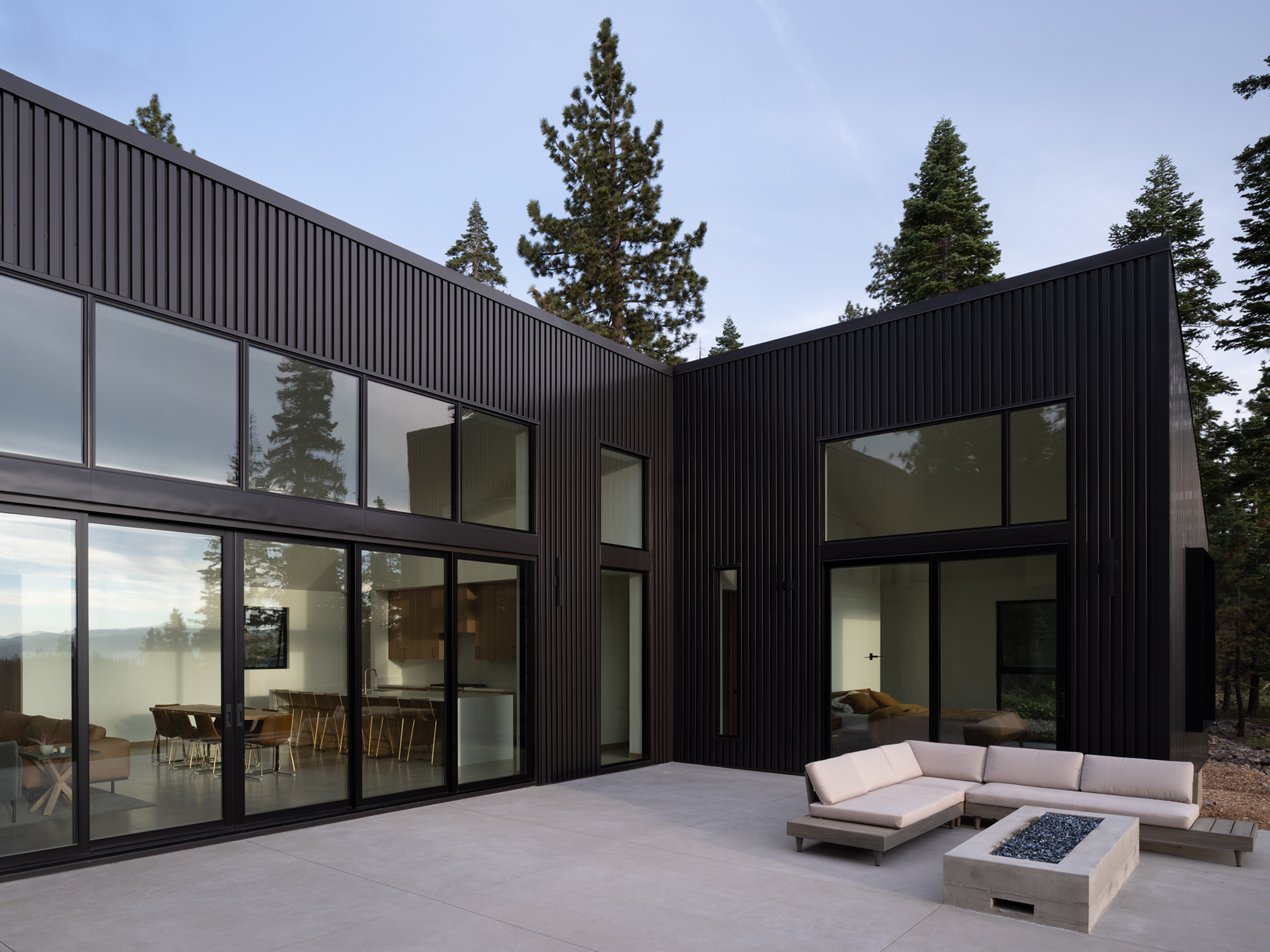 Modern black-clad house with large glass windows, outdoor patio with white sectional sofa and rectangular fire pit, surrounded by trees.