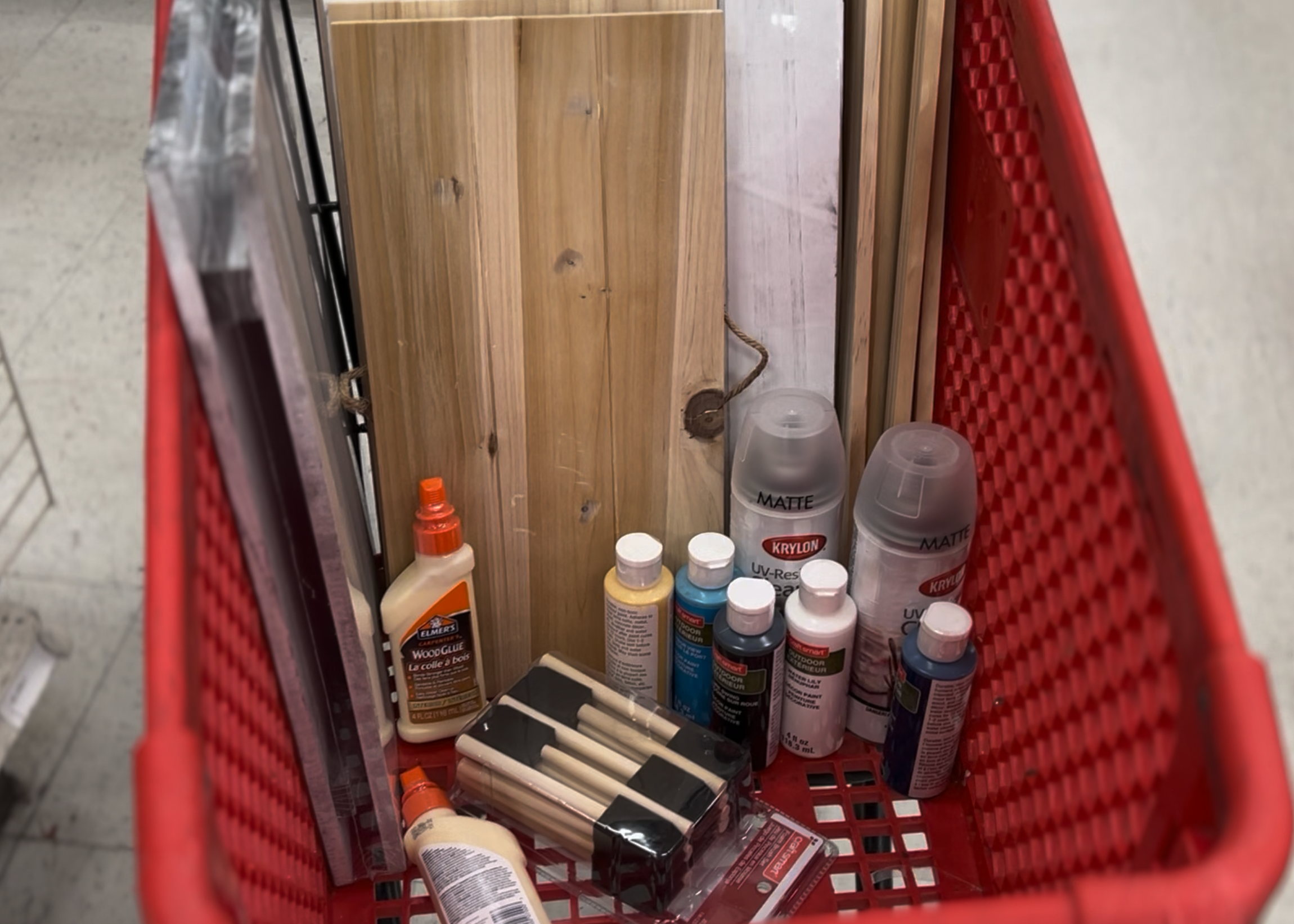 Inside a red shopping basket containing various bottles of spray paint, glue, and other craft supplies, with a wooden panel in the background.