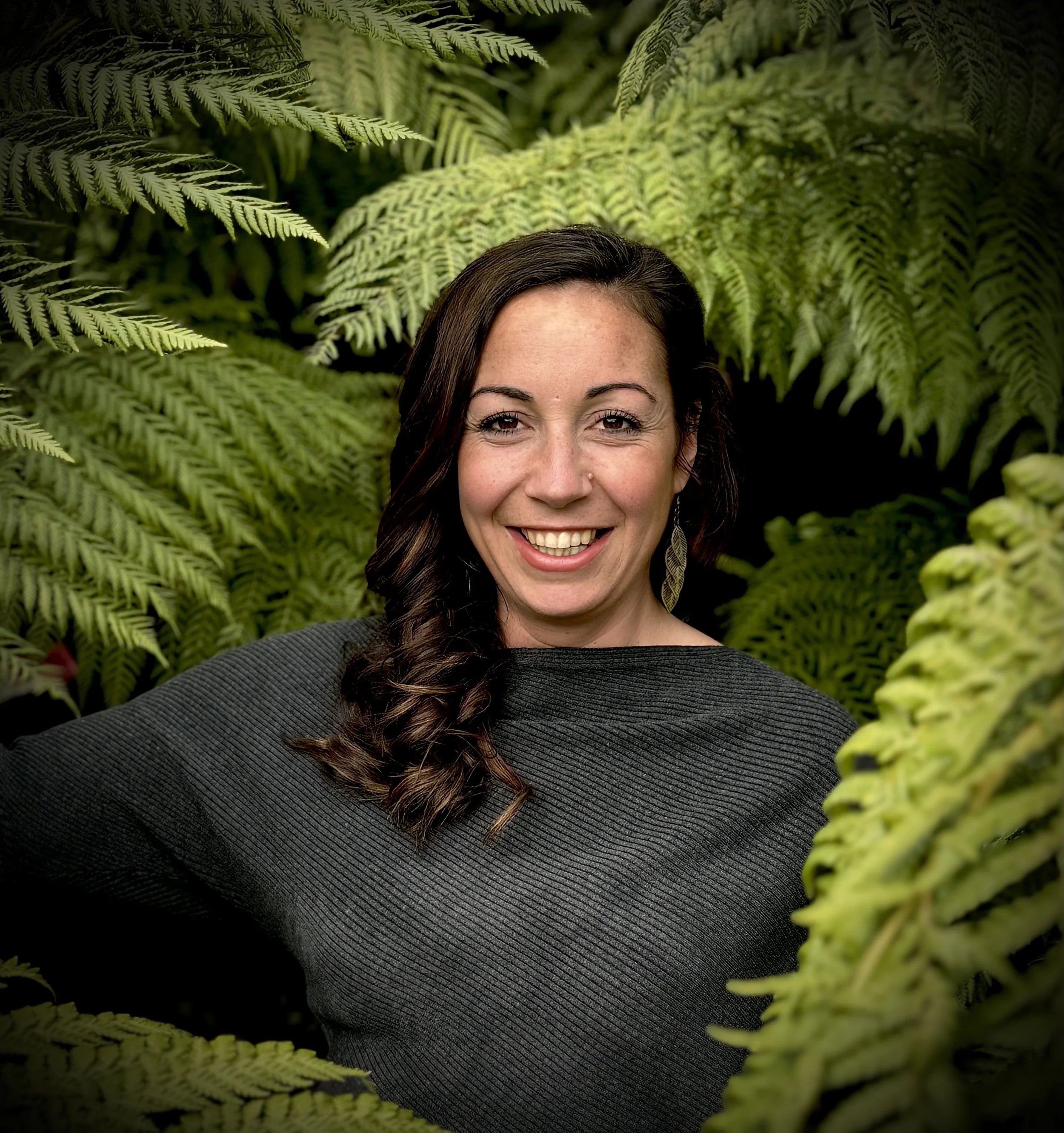 Part of a smiling woman's face with dark brown hair, surrounded by lush green fern leaves.