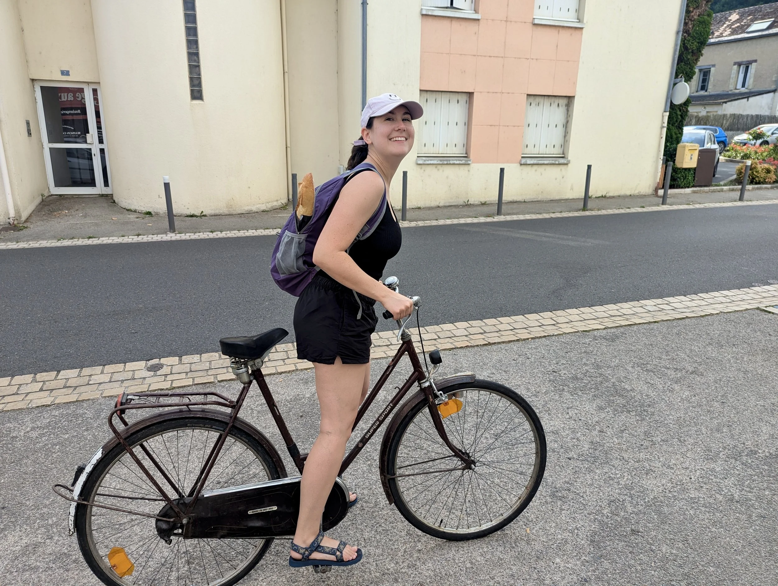 A woman smiling and wearing a white cap, black tank top, and shorts, is standing outdoors on a sidewalk with a bicycle, with buildings and parked cars in the background.