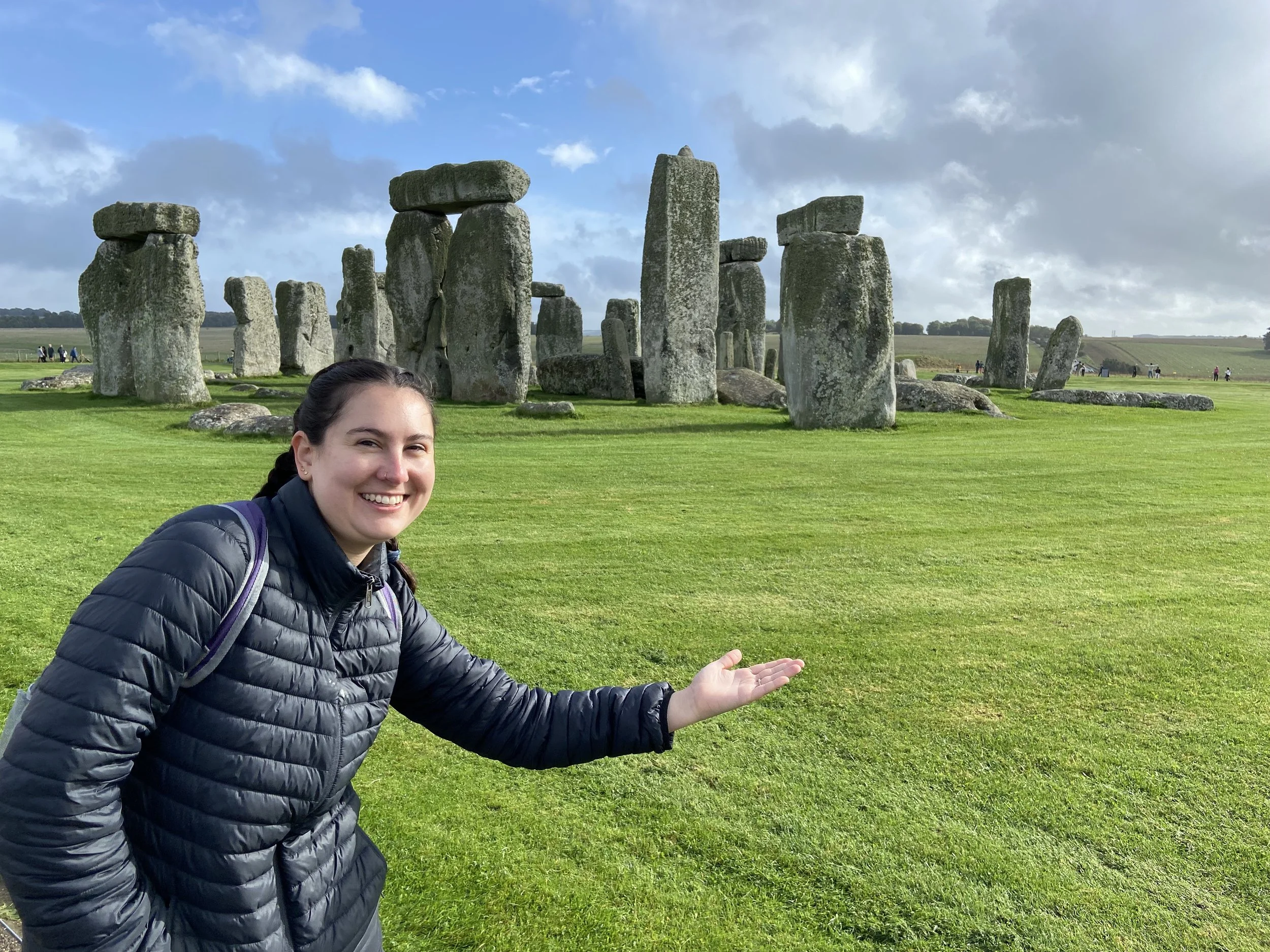 A smiling woman with dark hair in a braid, wearing a black puffy jacket, stands in front of the ancient stone monument of Stonehenge on a bright day with partly cloudy skies.