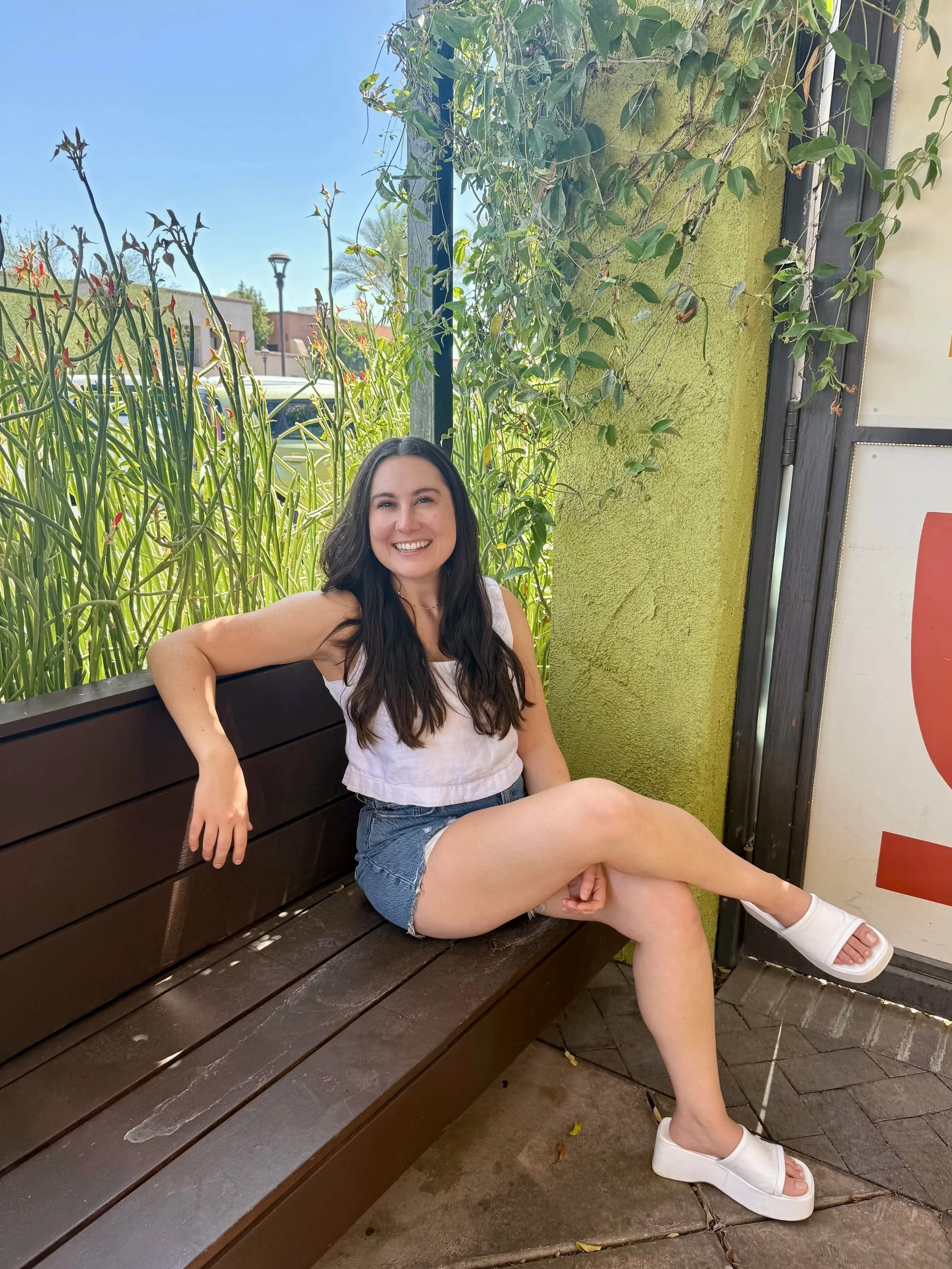 A young woman with long dark hair smiling and sitting on a wooden bench outdoors. She is wearing a light-colored sleeveless top, denim shorts, and white platform sandals. She is posing with her left arm resting on the back of the bench and her right leg crossed over her left, with a green wall, plants, a lamppost, and a street visible in the background.