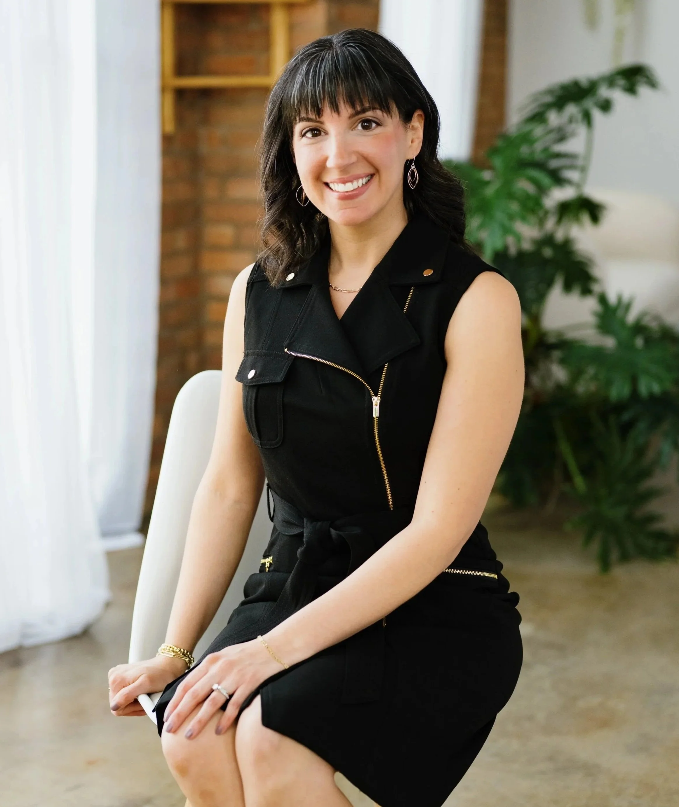 Woman sitting on chair, smiling, wearing a black sleeveless dress, in an indoor setting with a plant in the background
