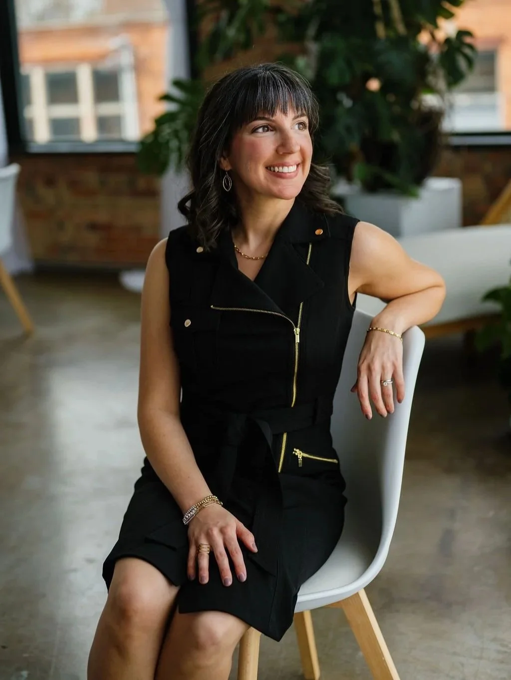 A woman with dark hair and bangs smiling while sitting on a white chair in a well-lit room with large windows and indoor plants.