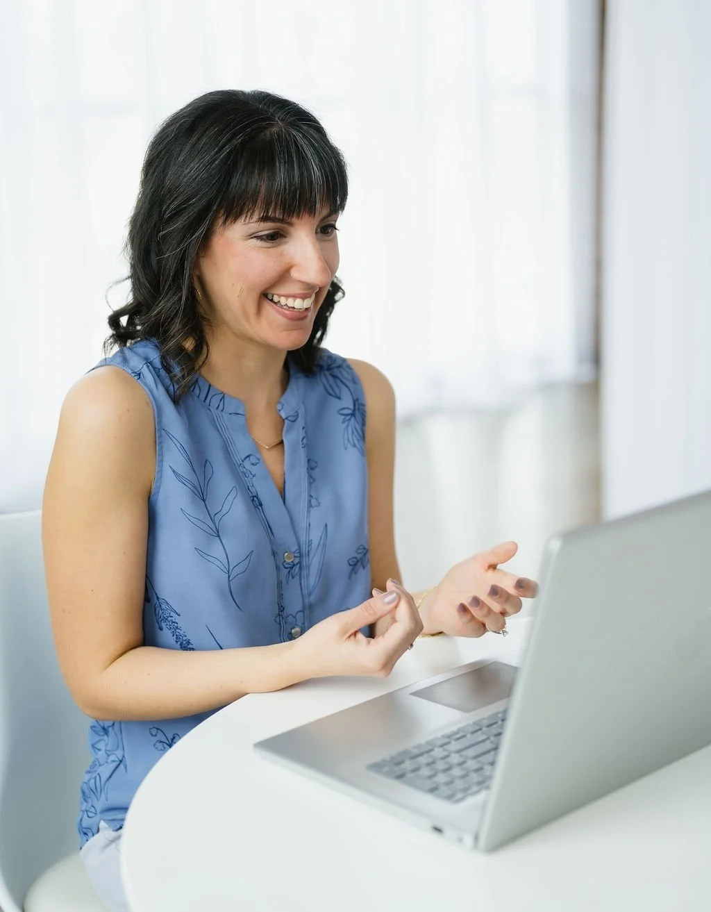 A woman with dark hair wearing a blue sleeveless top sitting at a white table using a silver laptop, smiling and engaging with the screen.