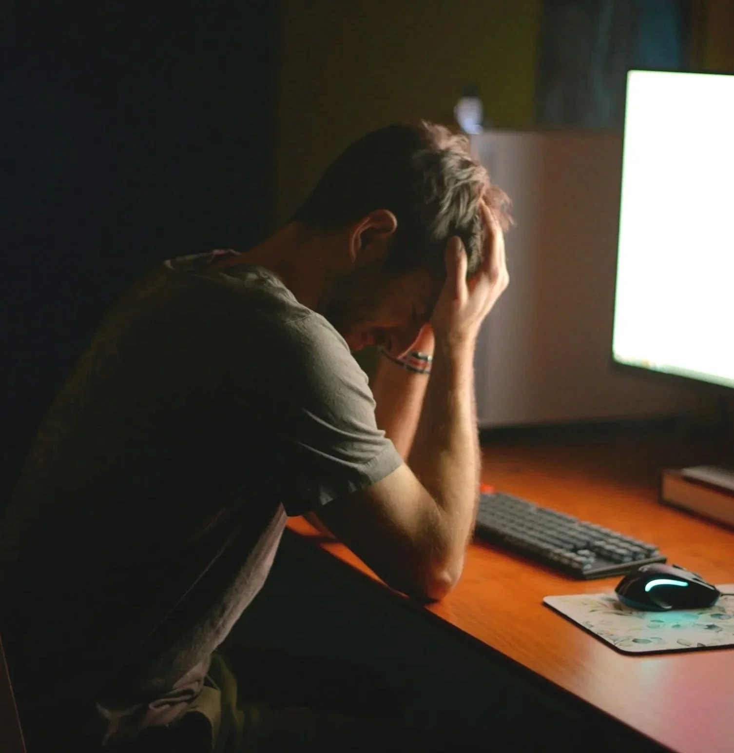 A young man sitting at a desk with his head in his hands, appearing stressed or frustrated while facing a computer monitor.