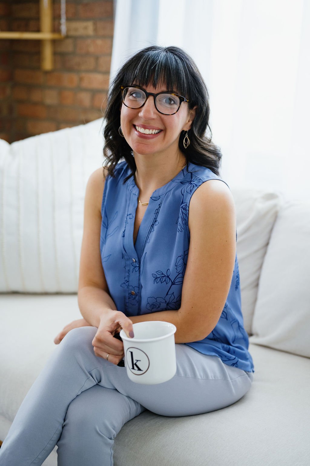 A woman with dark hair and glasses sitting on a beige sofa, holding a white mug with a black letter 'K', smiling at the camera.