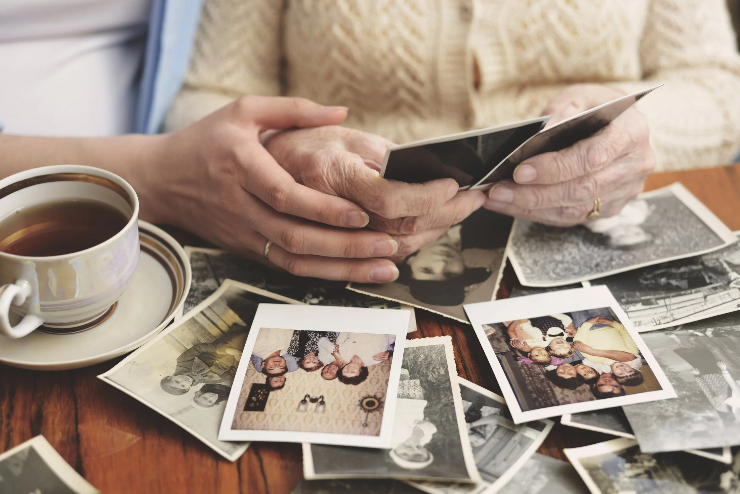 Older person and younger person looking at photo album with old photographs and printed photos, with a cup of tea on a wooden table.