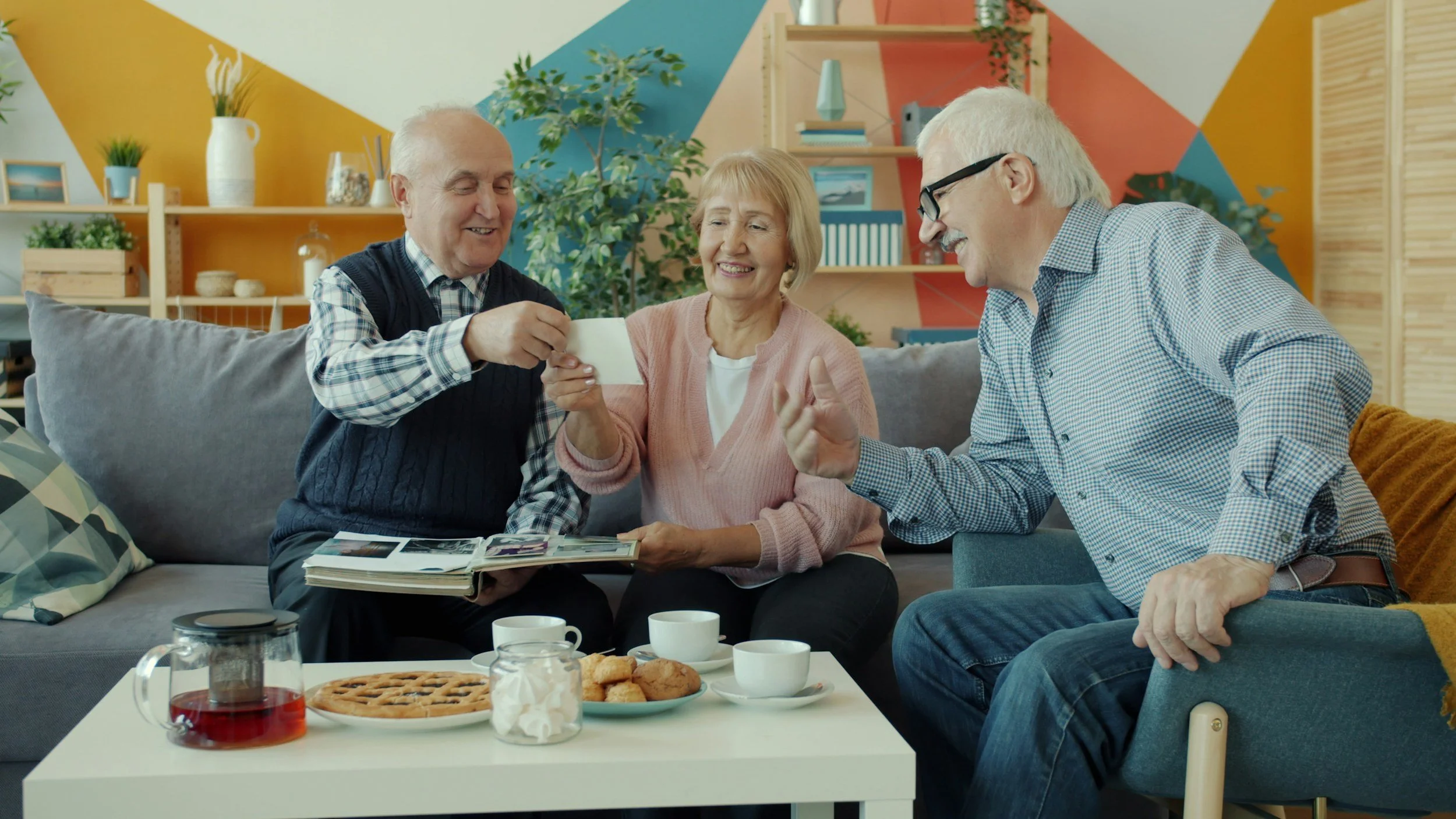 Three elderly people sitting on a sofa enjoying tea, cookies, and sharing photographs in a cozy, colorfully decorated living room.