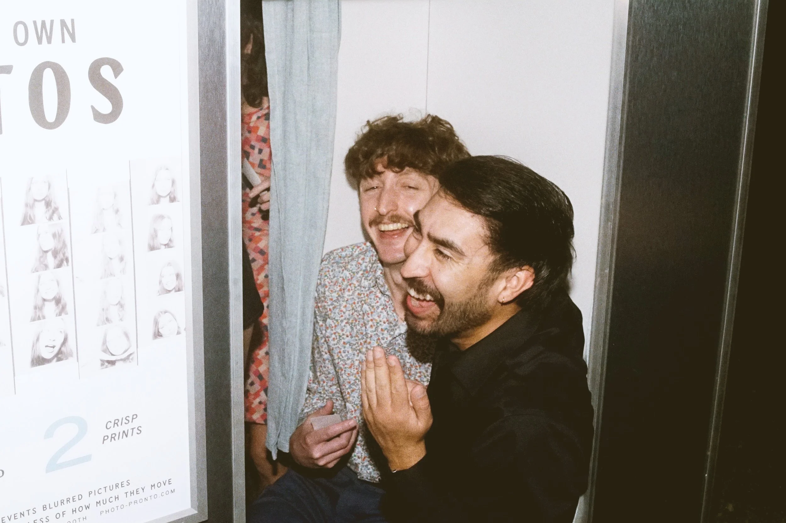Two men laughing happily in a vintage photo booth in Austin, one holding a photo booth strip, with a photo booth sign nearby.