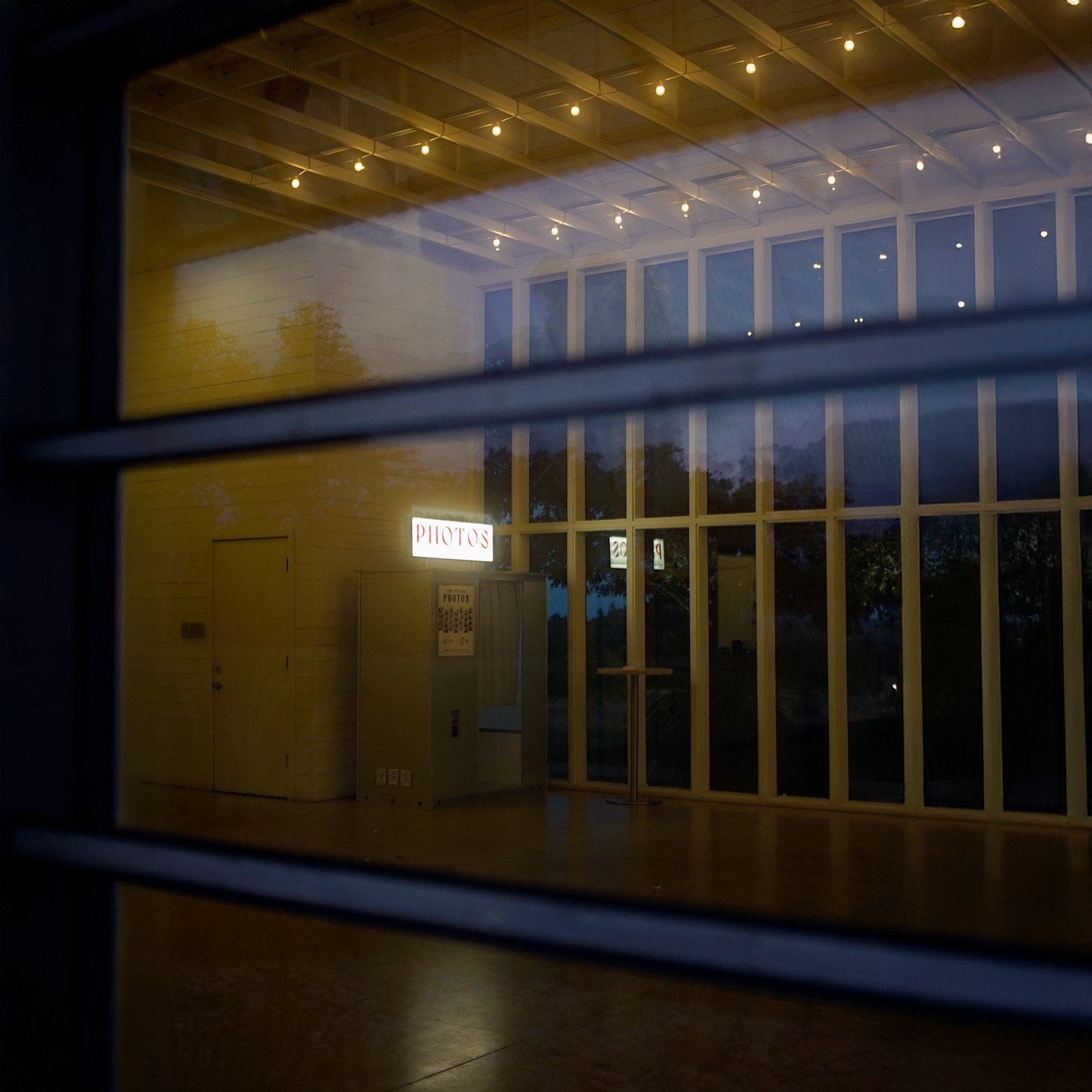 View through window showing a wedding venue with string lights on the ceiling, a classic photo booth, and views of trees outside.