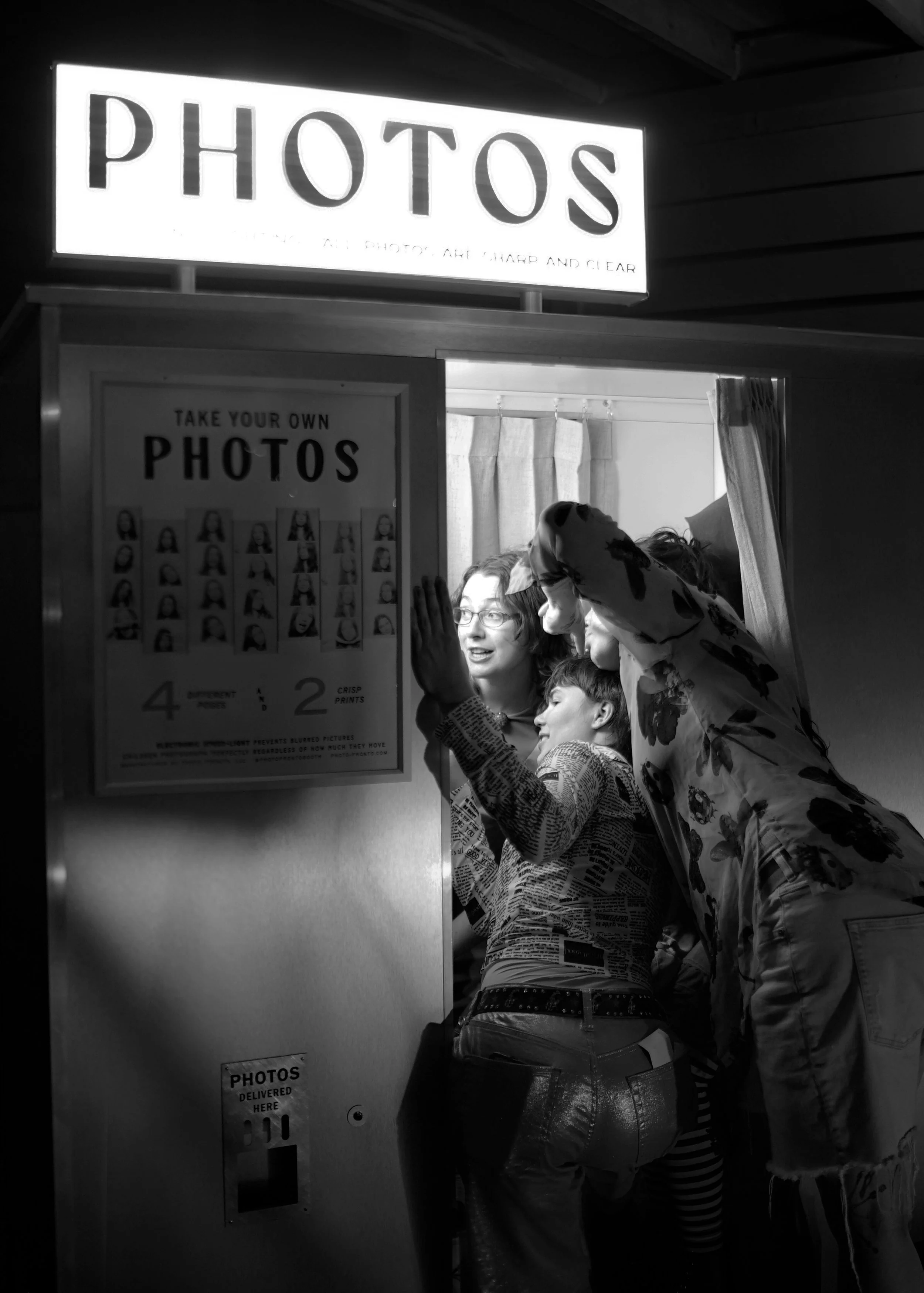 A black and white photo of a photo booth with three women, one with glasses, taking a picture together.