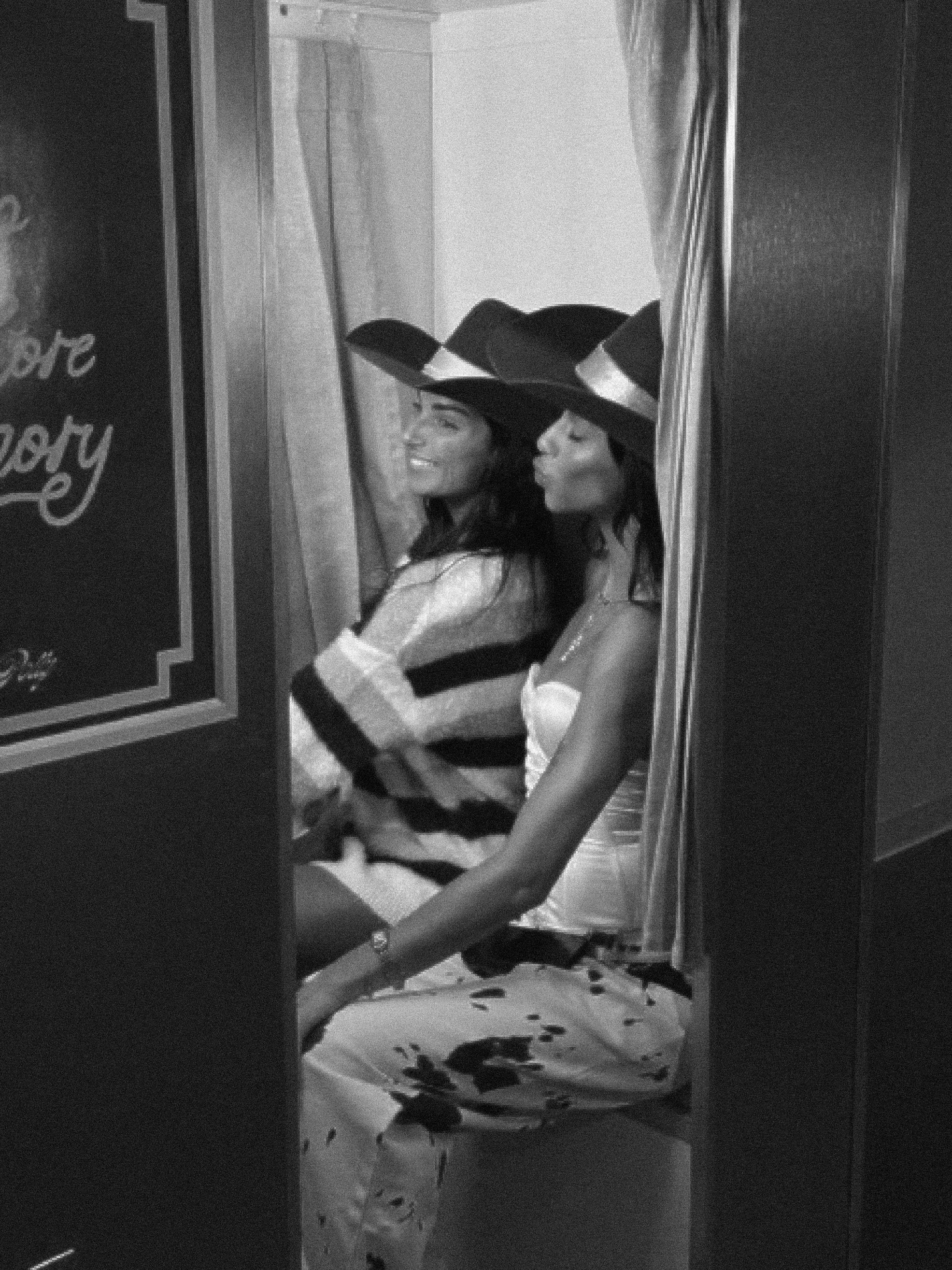Two women sitting together, both wearing cowboy hats, one smiling and the other puckering her lips, in a photo booth with curtain. 